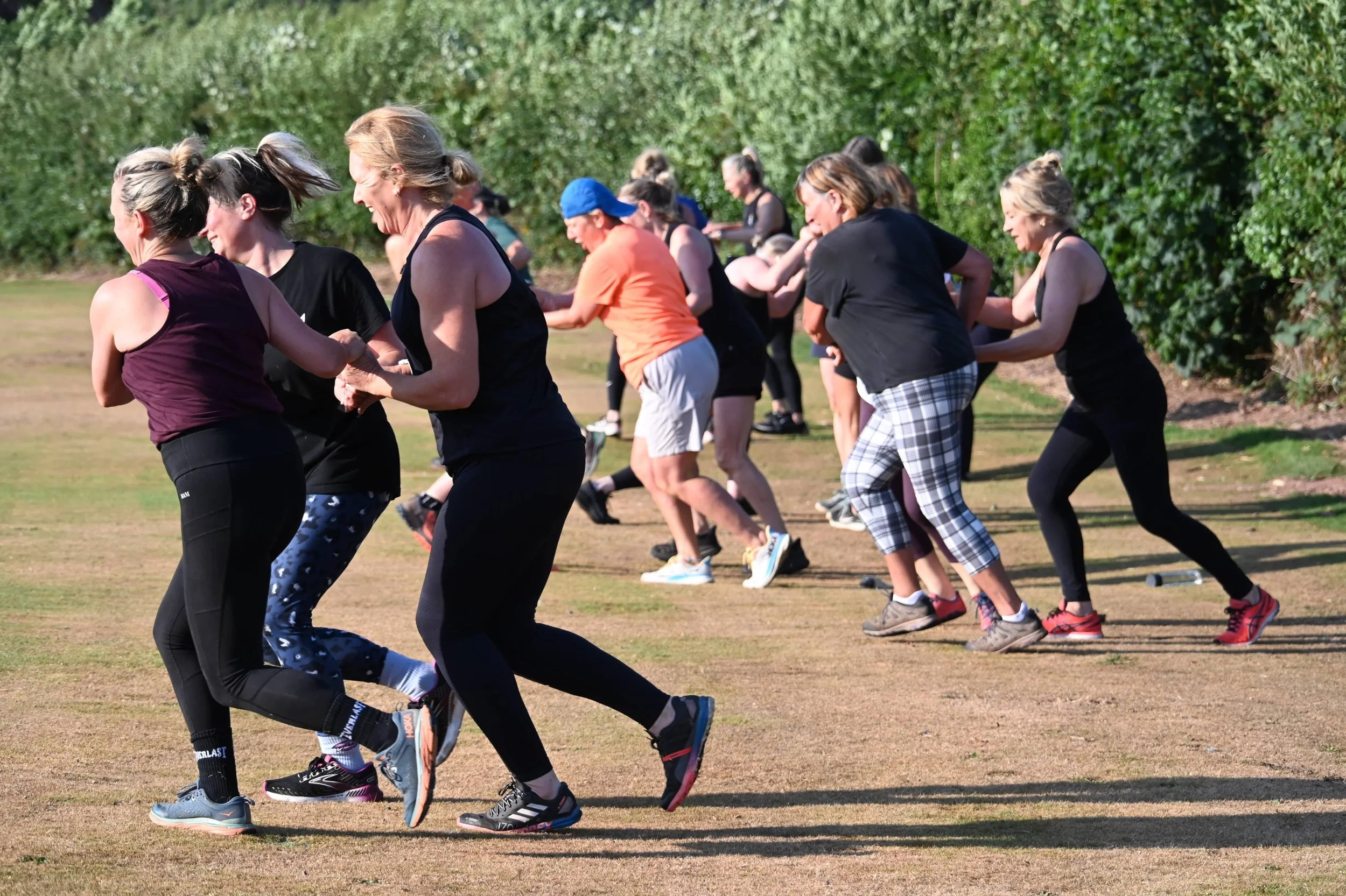A group of women running and participating in an outdoor exercise class on a grassy field, with a background of green bushes and trees.