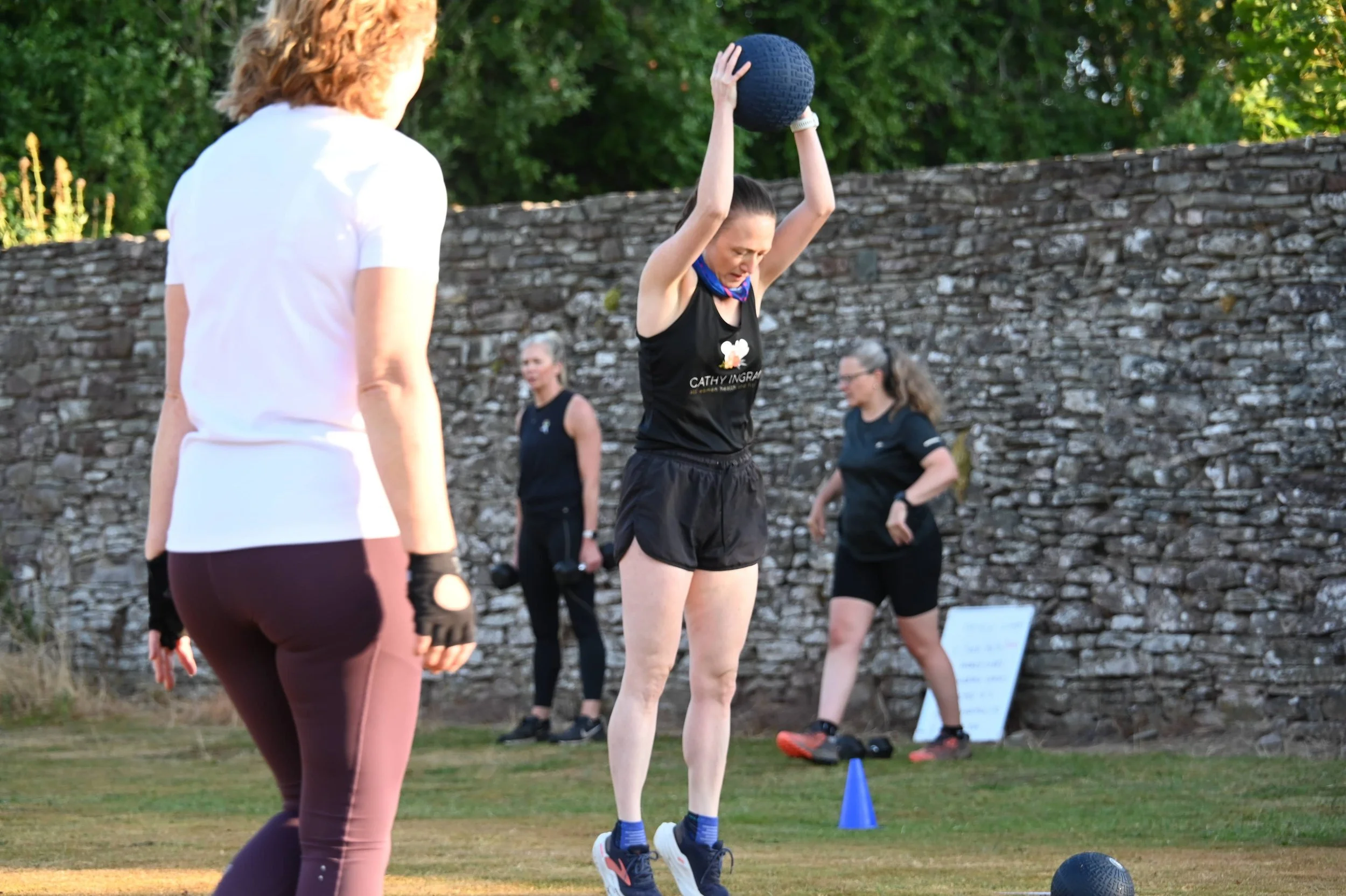 Women participating in an outdoor fitness class, one lifting a medicine ball overhead, others standing around with fitness equipment, on a grassy area with a stone wall and green trees in the background.