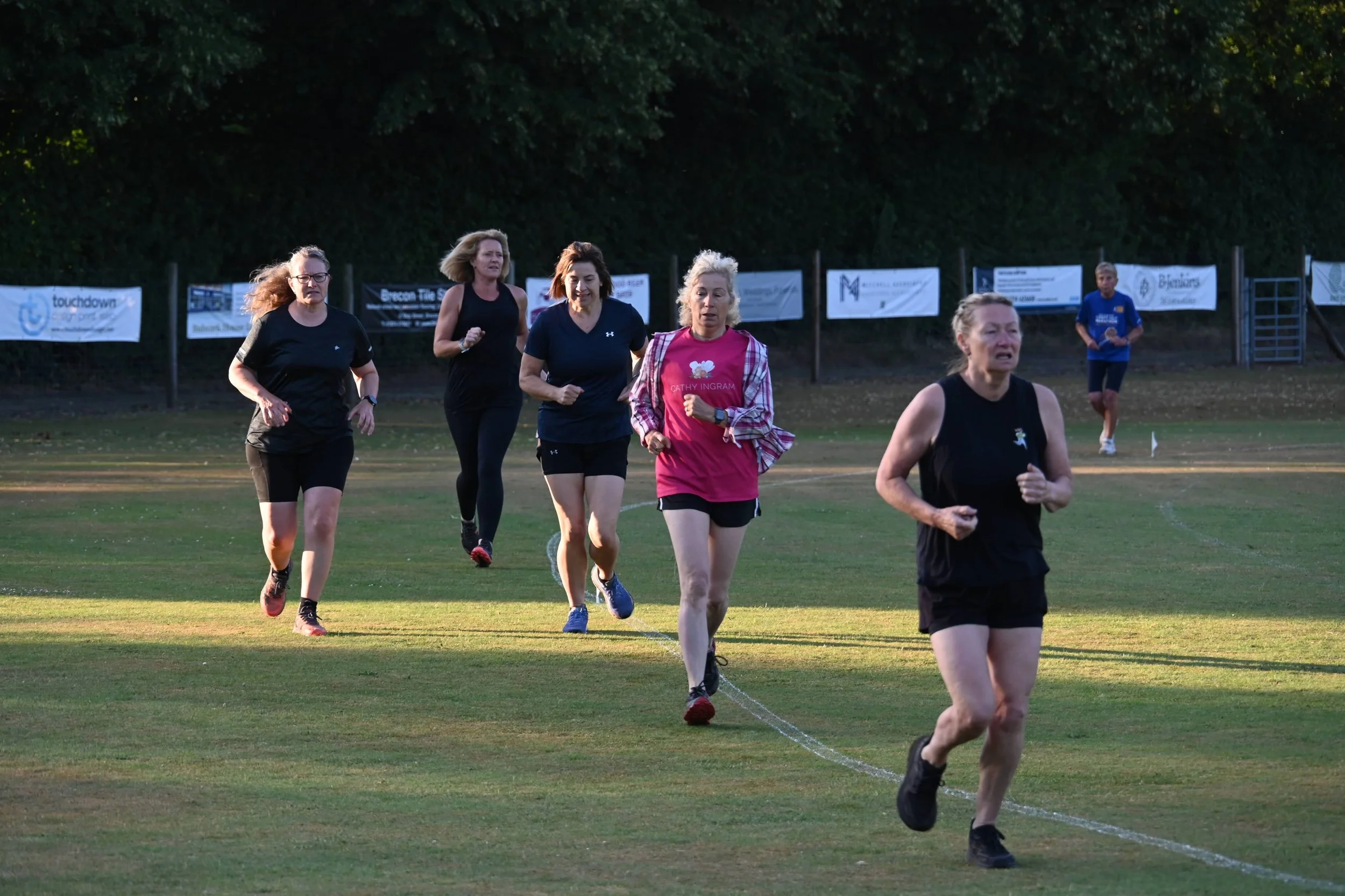 A group of women running on a grass field during a race or event, with a backdrop of trees and banners in the background.