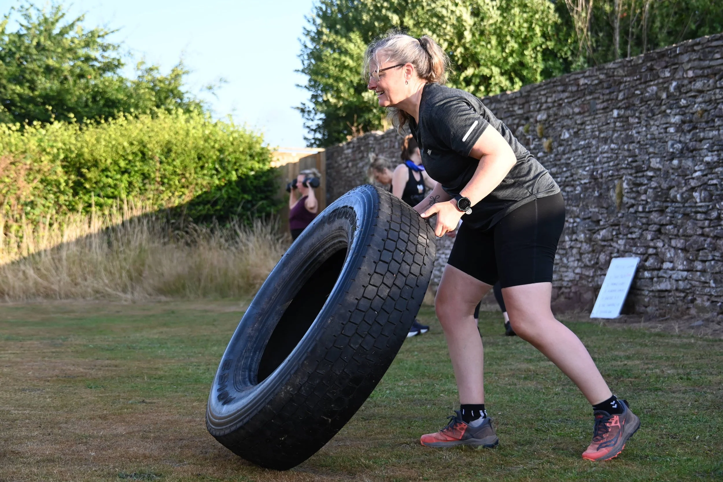 A woman with glasses and a watch lifting a large black tire outdoors during daytime, with two women in the background near a stone wall and trees.