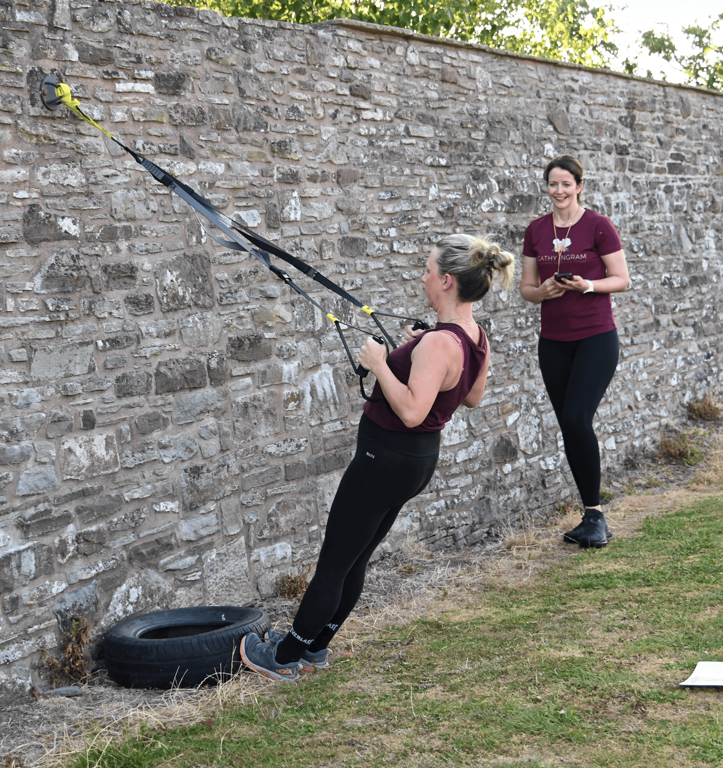 Two women exercising outdoors against a stone wall, with one woman using suspension training equipment anchored to the wall and the other woman holding a smartphone and smiling.