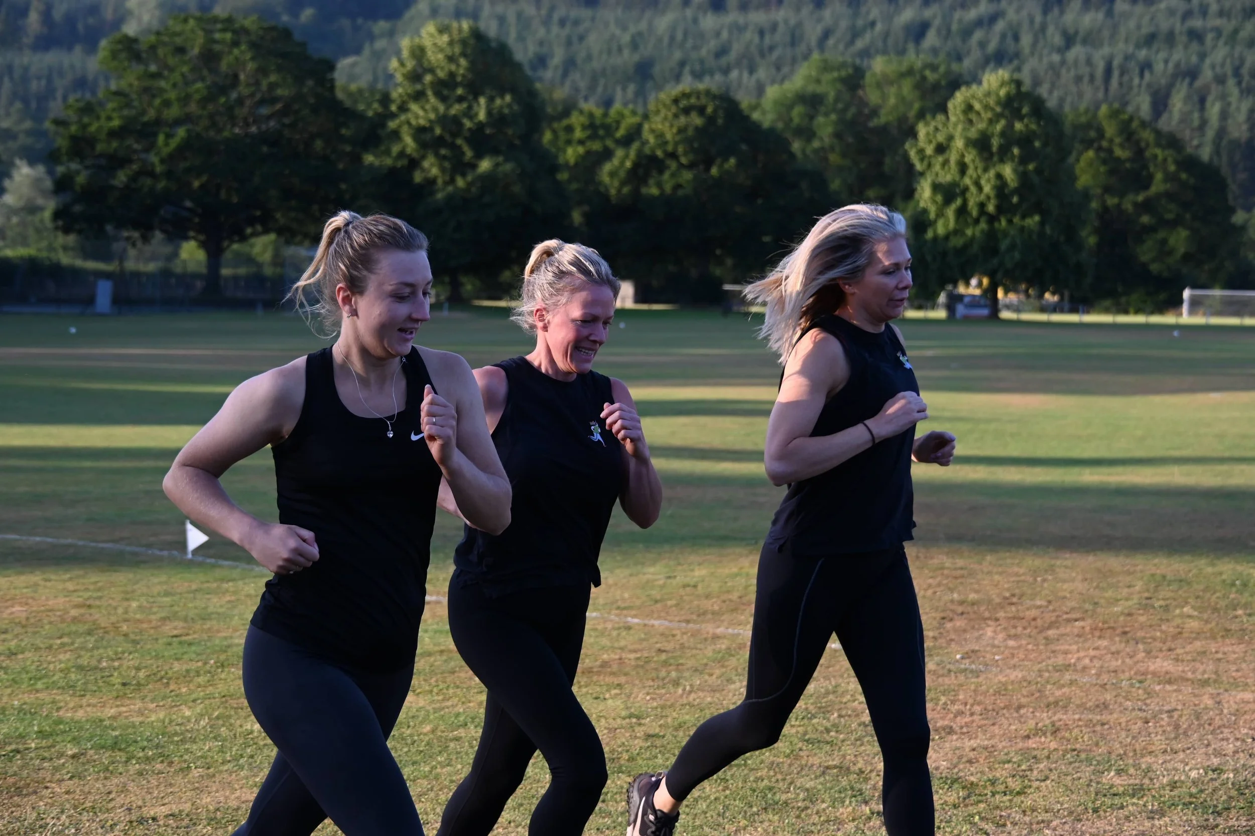 Three women running on a grassy field in an outdoor park with trees and mountains in the background.