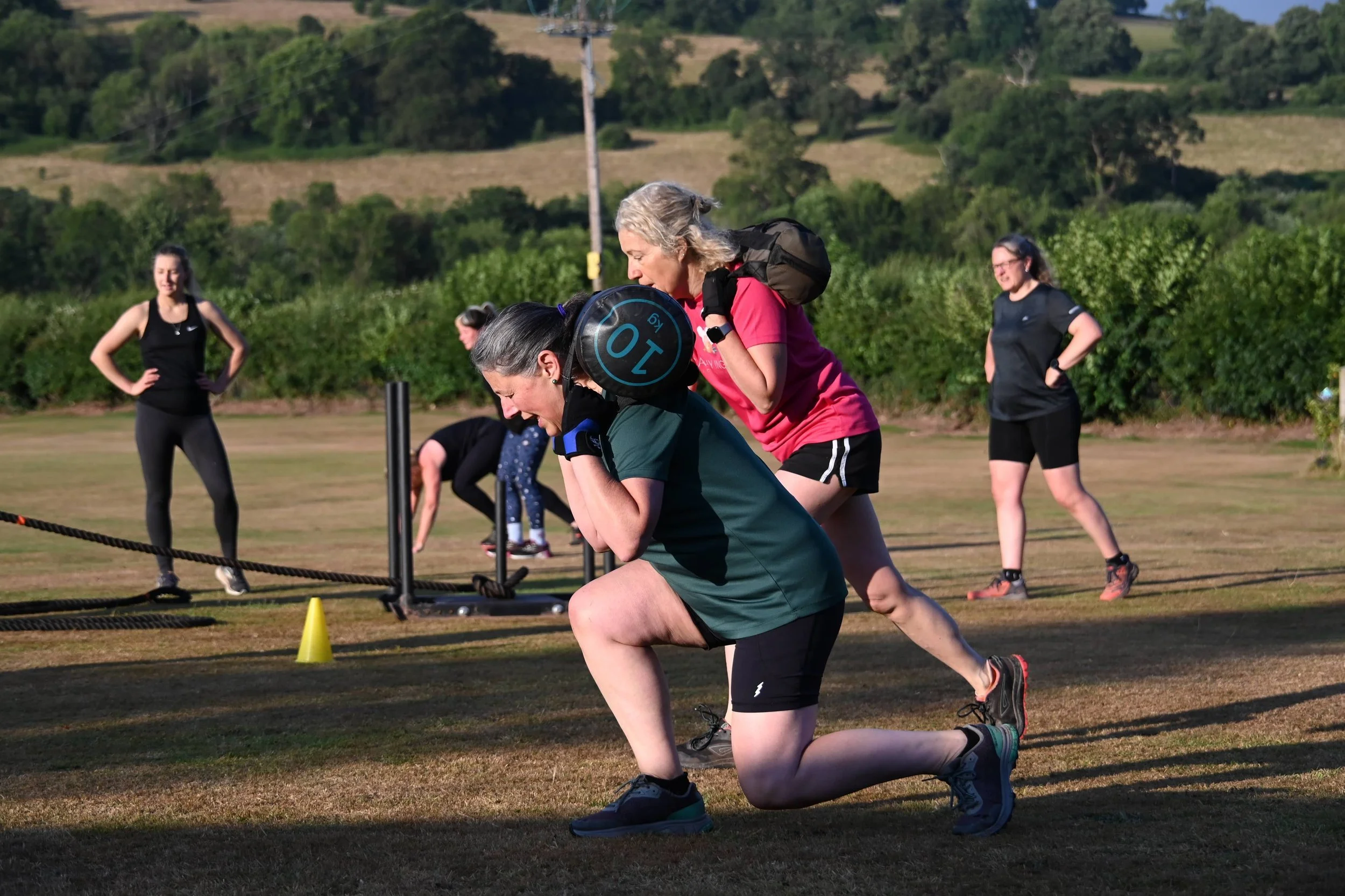 Women participating in an outdoor fitness class, kneeling and bending forward with exercise equipment on their backs, on a grassy field with hills and trees in the background.