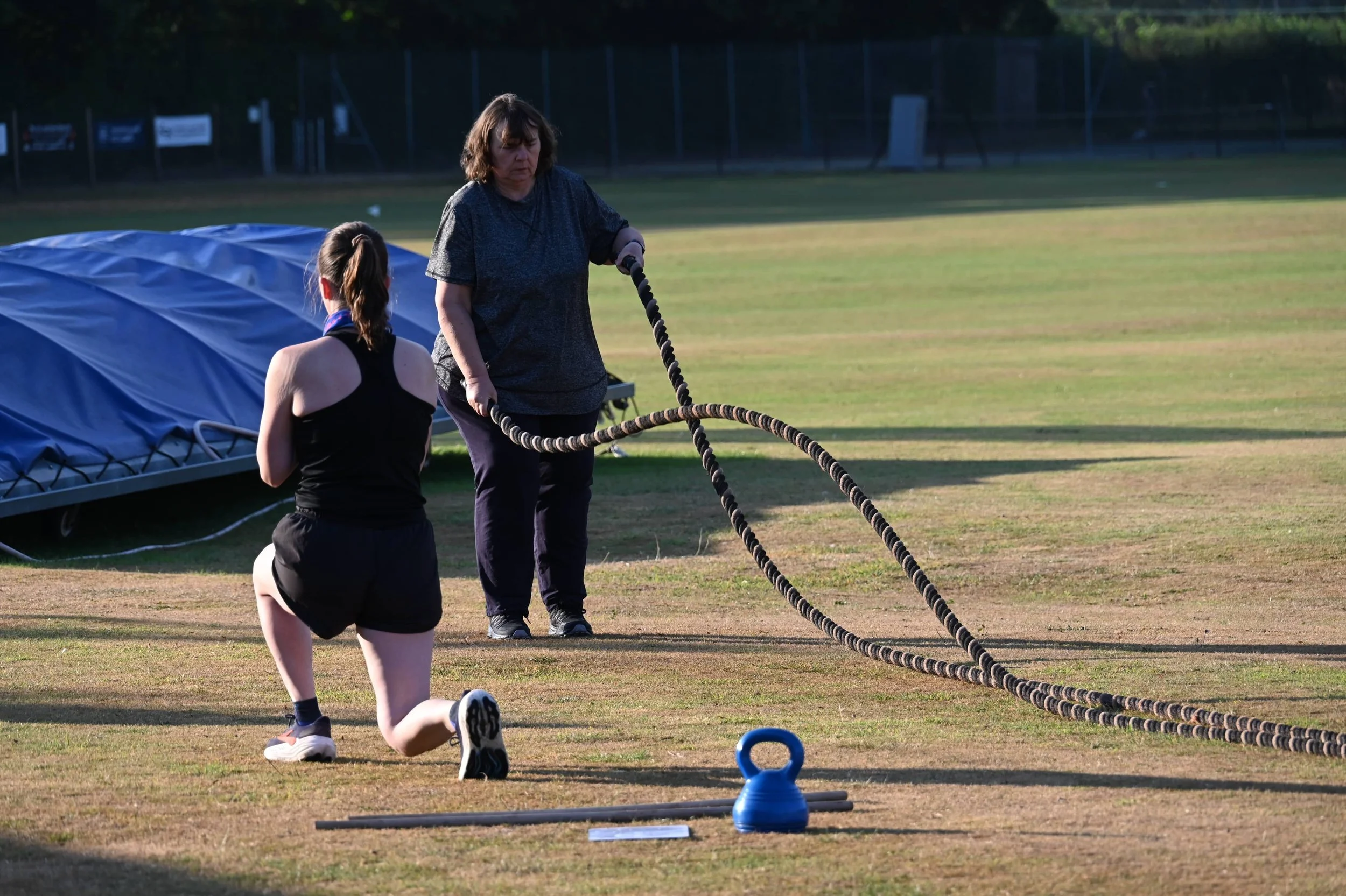 A woman and a girl are exercising outdoor on a field with fitness equipment, including ropes and a kettlebell.