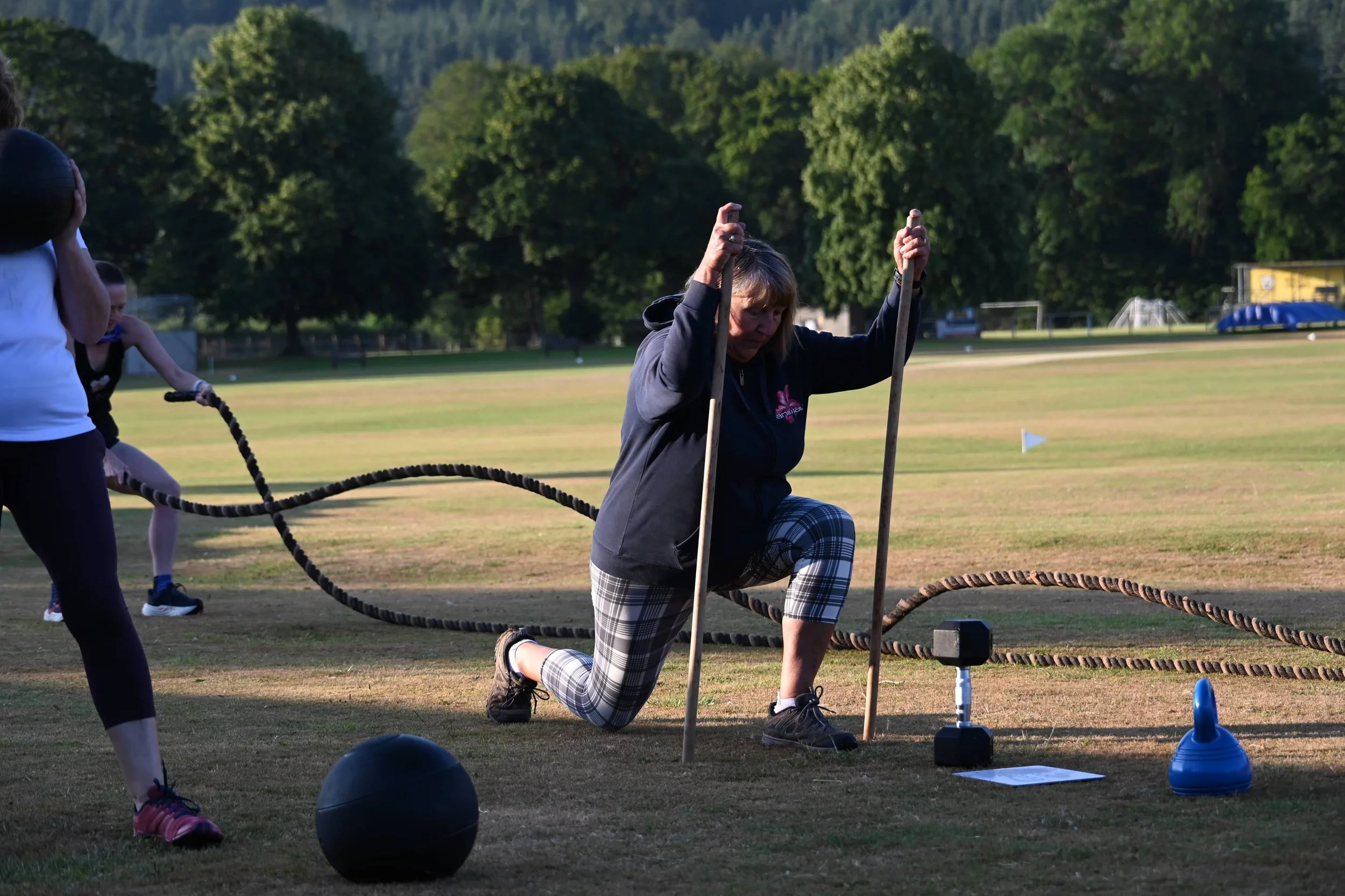 A woman is kneeling on one knee, holding a wooden ring with both hands, participating in an outdoor activity on a field. Other people are also involved, and there are sports cones and equipment around, with trees and hills in the background.