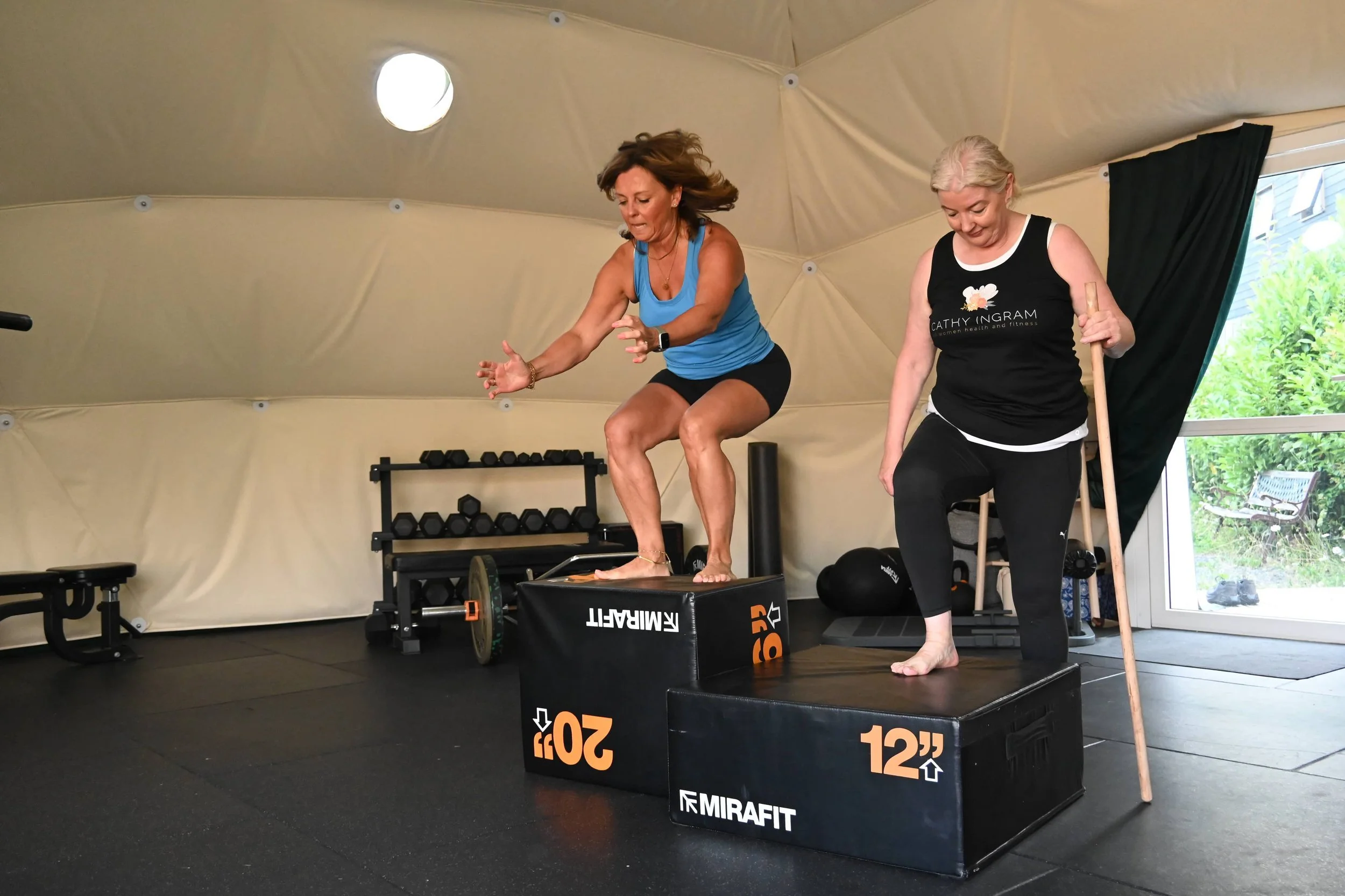 Two women exercising in an indoor gym. One woman is jumping onto a plyometric box, while the other is holding a stick and observing. The gym has weights, kettlebells, and workout equipment, with a large window showing outdoor greenery.