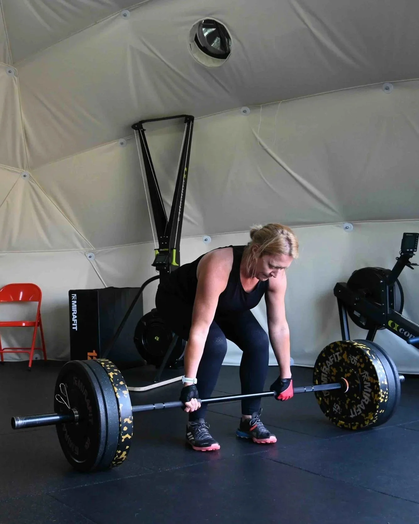 A woman lifting a heavy barbell during a workout in a fitness gym, with exercise equipment around her and a white enclosure in the background.