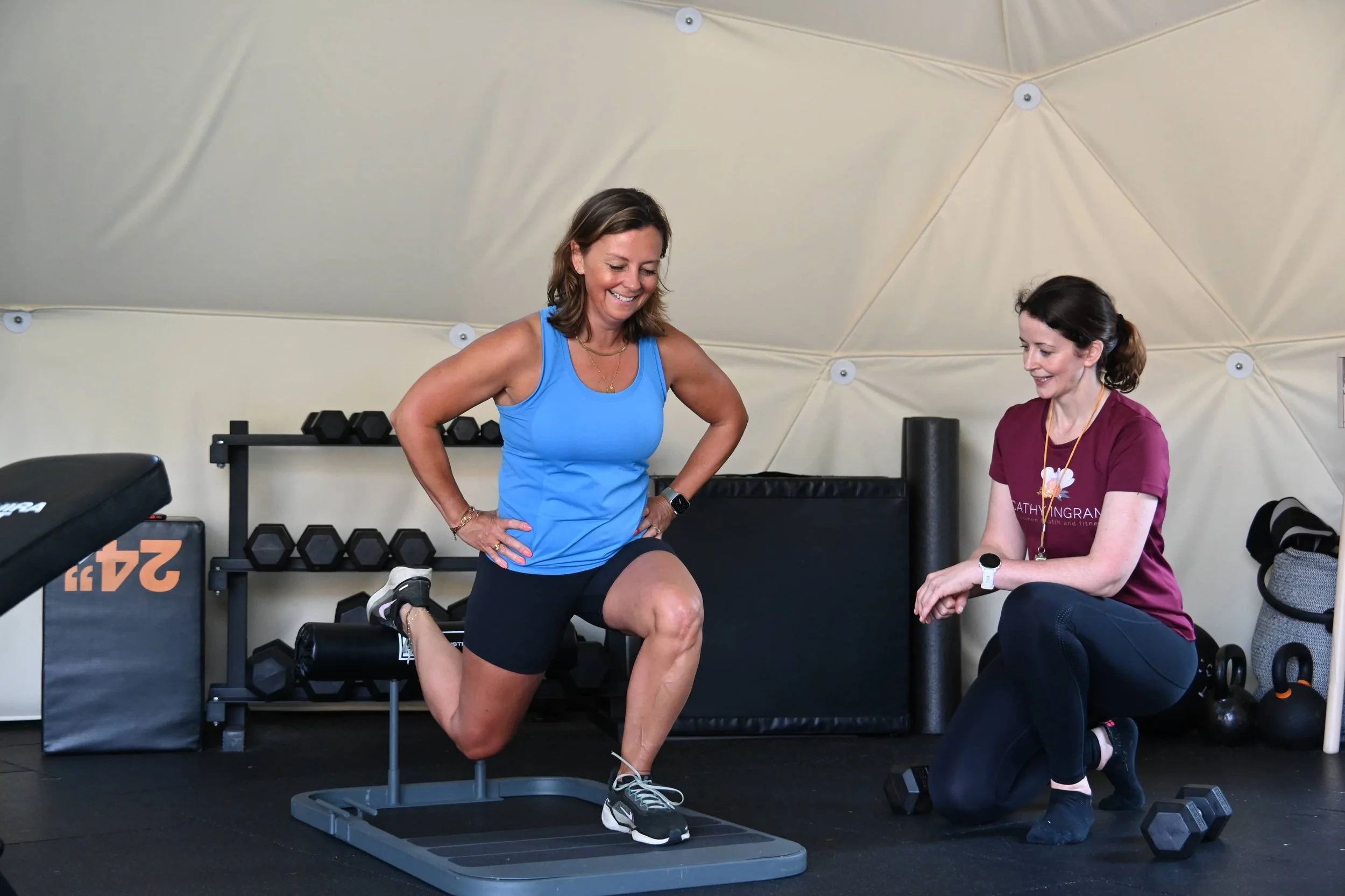 Two women in a gym. One woman in a blue tank top and black shorts is performing a lunge exercise on a step platform, smiling and looking down at her knee. The other woman in a maroon shirt and black leggings is sitting beside her, providing support or coaching. Gym equipment, such as dumbbells and foam rollers, are visible in the background.