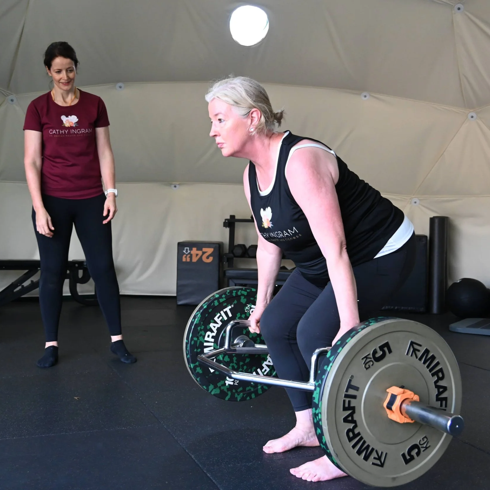 An elderly woman with gray hair lifting a weighted barbell during a strength training session at a gym, supervised by a trainer in a maroon shirt with 'Cathy Ingram' written on it.