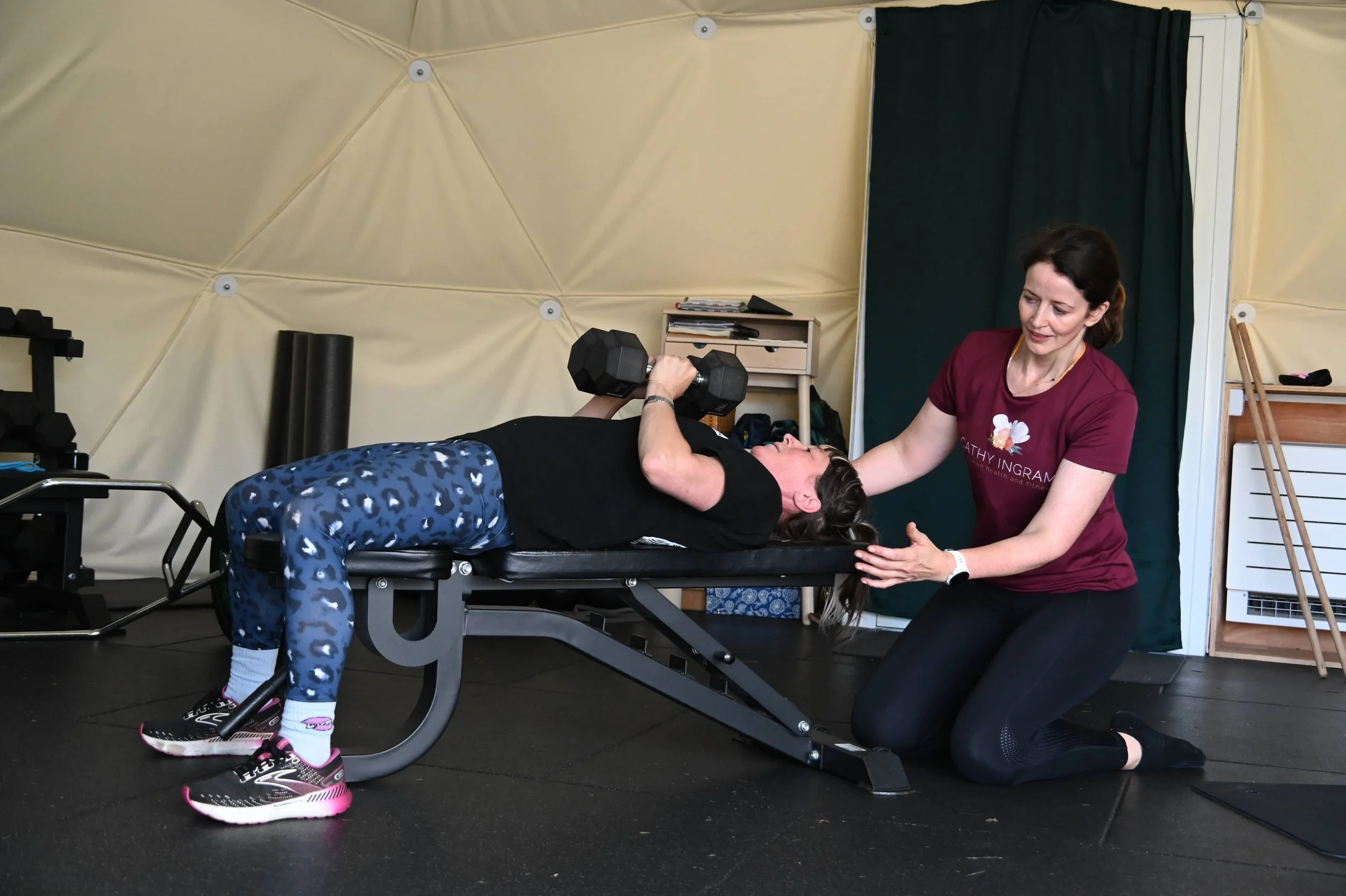 A woman in a maroon shirt assisting a woman lying on an exercise table while lifting a dumbbell. The scene appears to be in a fitness or rehabilitation setting.
