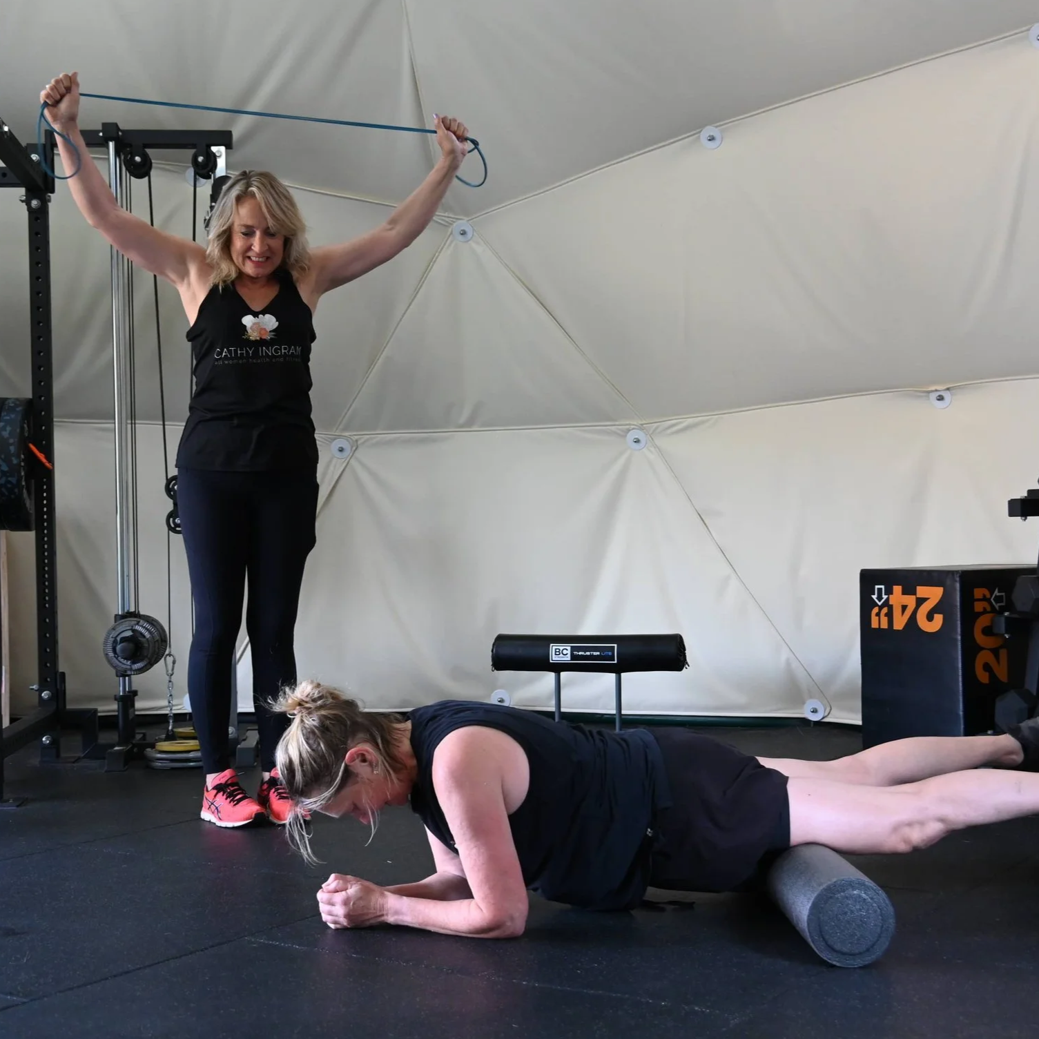 A woman is standing and holding a resistance band while another woman is doing a plank exercise in a gym.