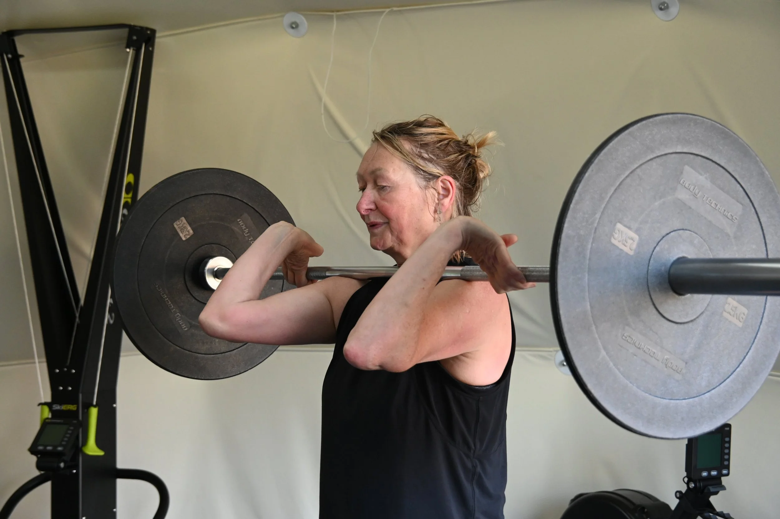 A woman with light skin and reddish-brown hair tied back, lifting a barbell during a workout in a gym.