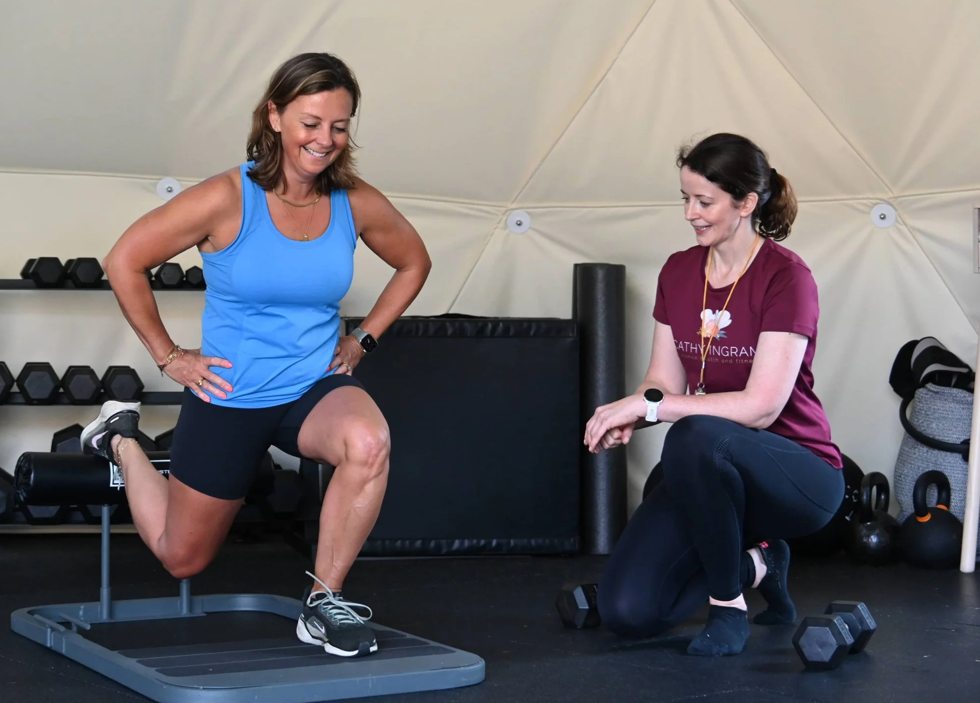Two women in a gym, one is exercising on a step platform while the other is sitting and observing. The woman exercising is wearing a blue tank top and black shorts, appears to be doing a knee lift exercise. The woman sitting is in a maroon t-shirt with the name 'Cathy Vingran' on it and black leggings. Gym equipment like dumbbells, a rolled-up mat, and weights are visible in the background.