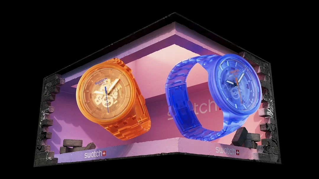 Two colorful Swatch watches, one orange and one blue, displayed in a glass enclosure against a pink background with broken wall edges.