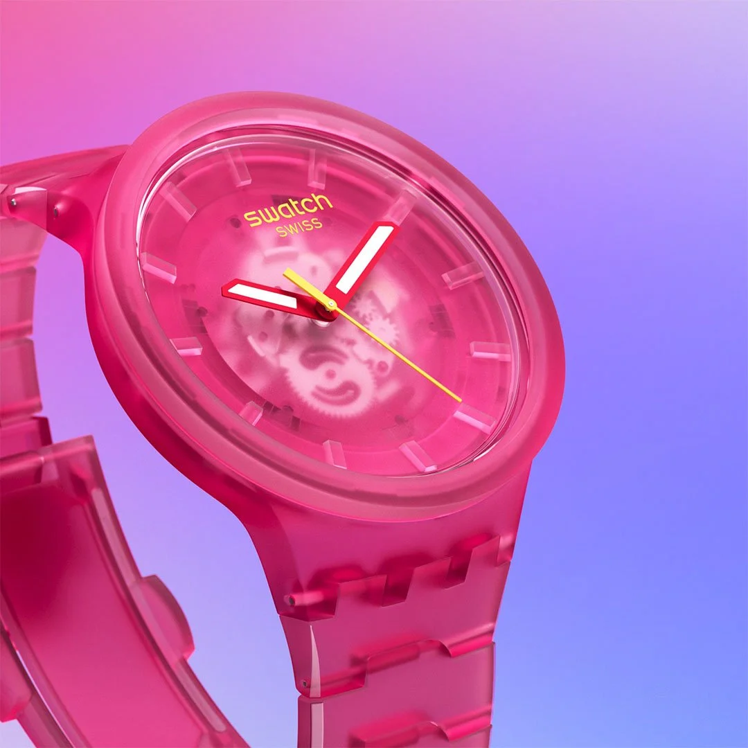 Close-up of a pink Swatch watch with a transparent face showing gears inside, set against a gradient background of pink, purple, and blue.