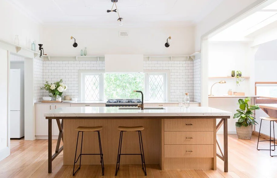 Bright, modern kitchen with white subway tile backsplash, wooden island with two stools, and large windows with greenery outside.