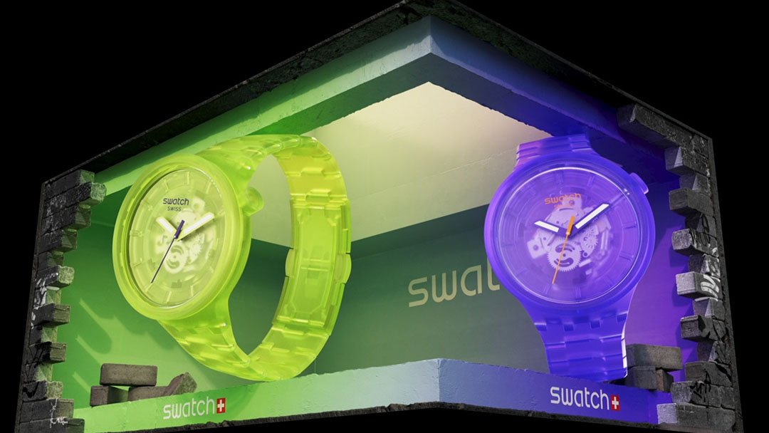 Display case featuring two colorful transparency Swatch watches, one yellow-green on the left and one purple on the right, inside a brick and foam display box.
