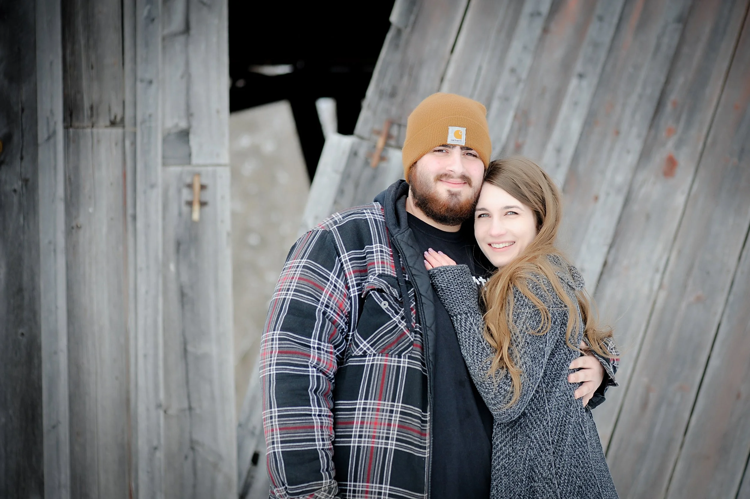A bearded man wearing a tan Carhartt beanie and a plaid jacket stands next to a smiling woman with long light brown hair, both hugging in front of a weathered wooden wall.