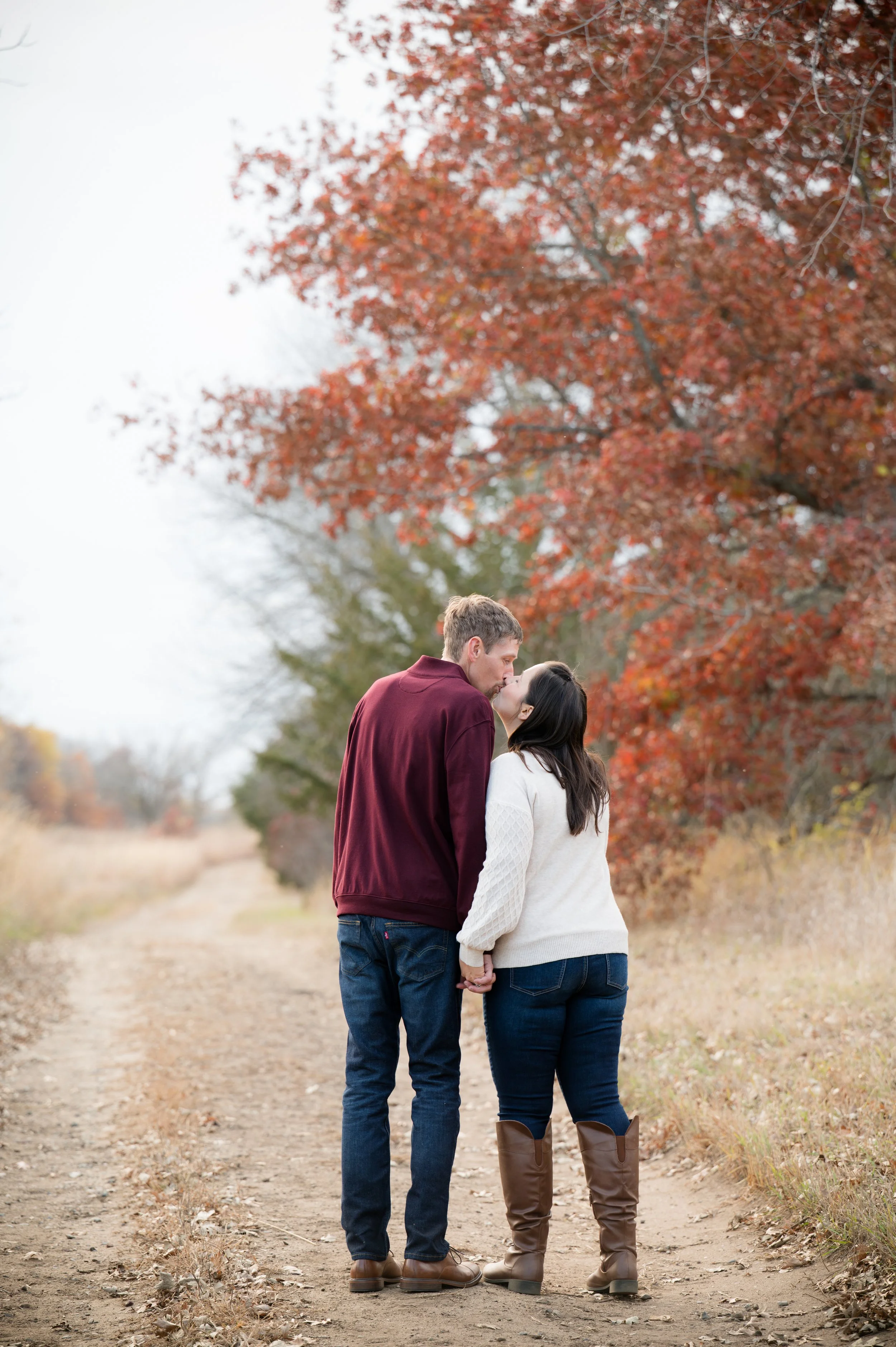 A couple holding hands and kissing on a dirt path in an autumn landscape with trees leaves in shades of red and orange.