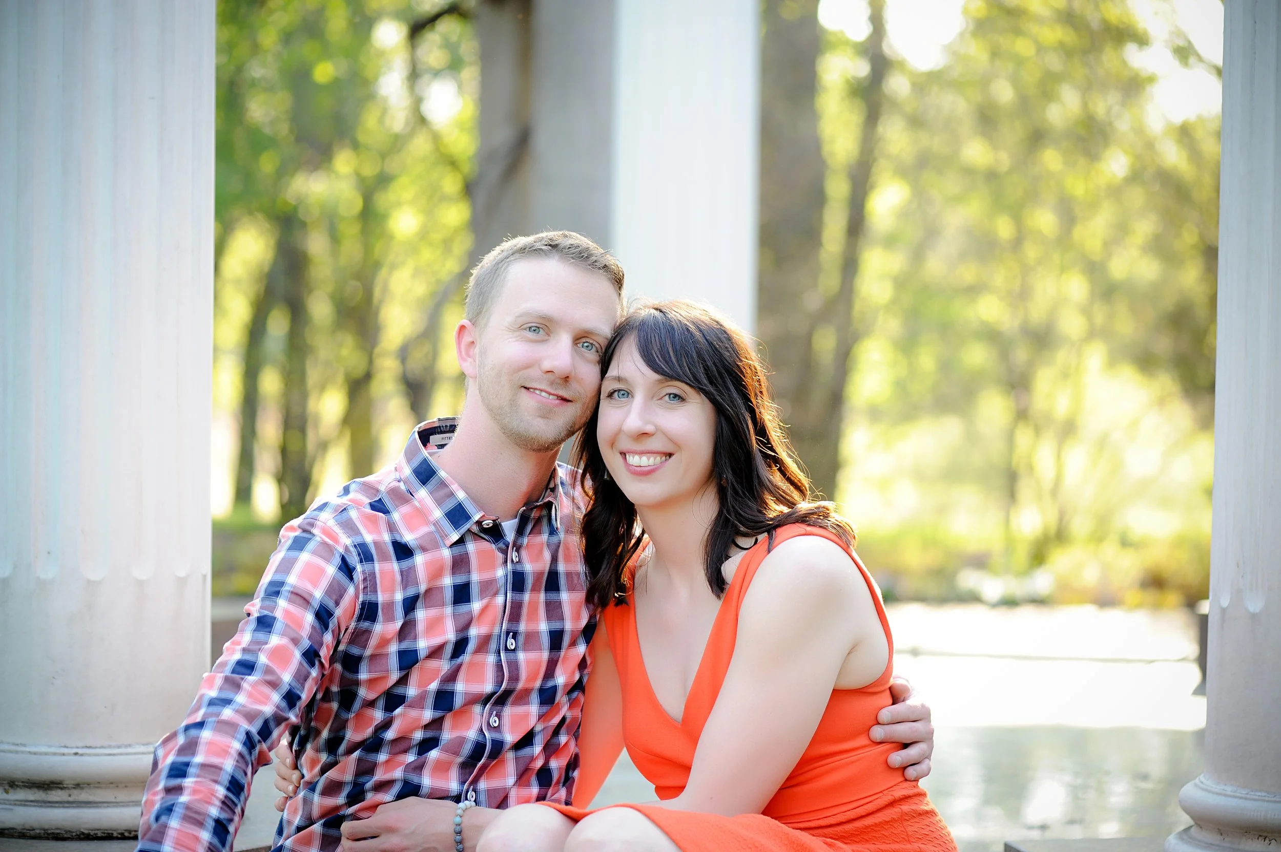 A young couple sitting together outdoors on a sunny day with trees in the background. The man has short blond hair and is wearing a plaid shirt, and the woman has long dark hair and is wearing an orange dress. They are smiling and hugging.