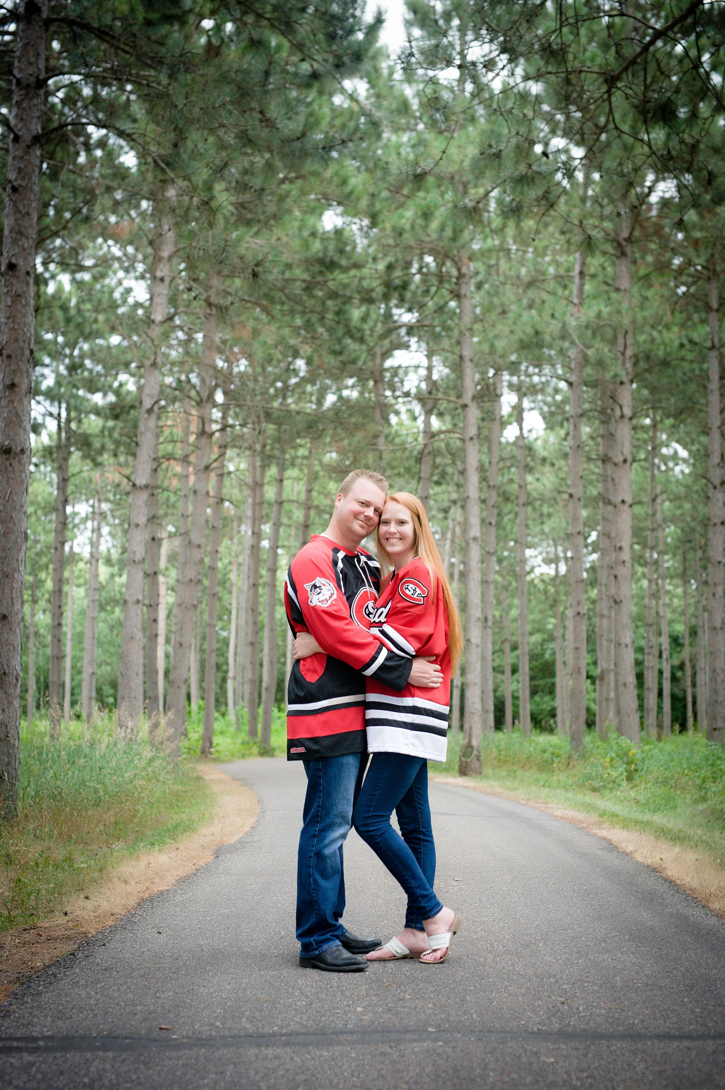 A couple wearing hockey jerseys standing on a tree-lined paved path in a forest, hugging and smiling at the camera.