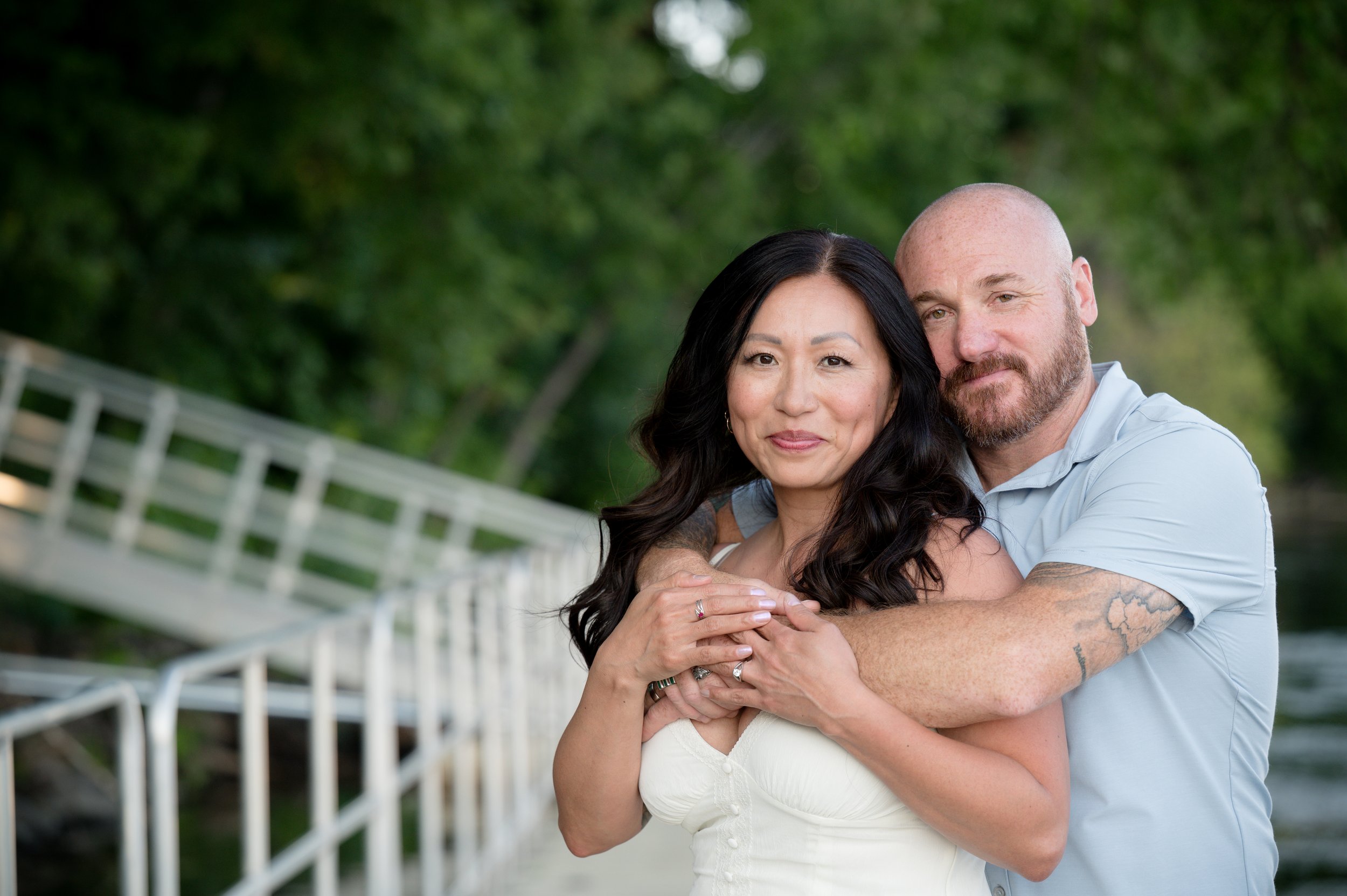 A couple embracing outdoors near a white railing and green trees in the background.