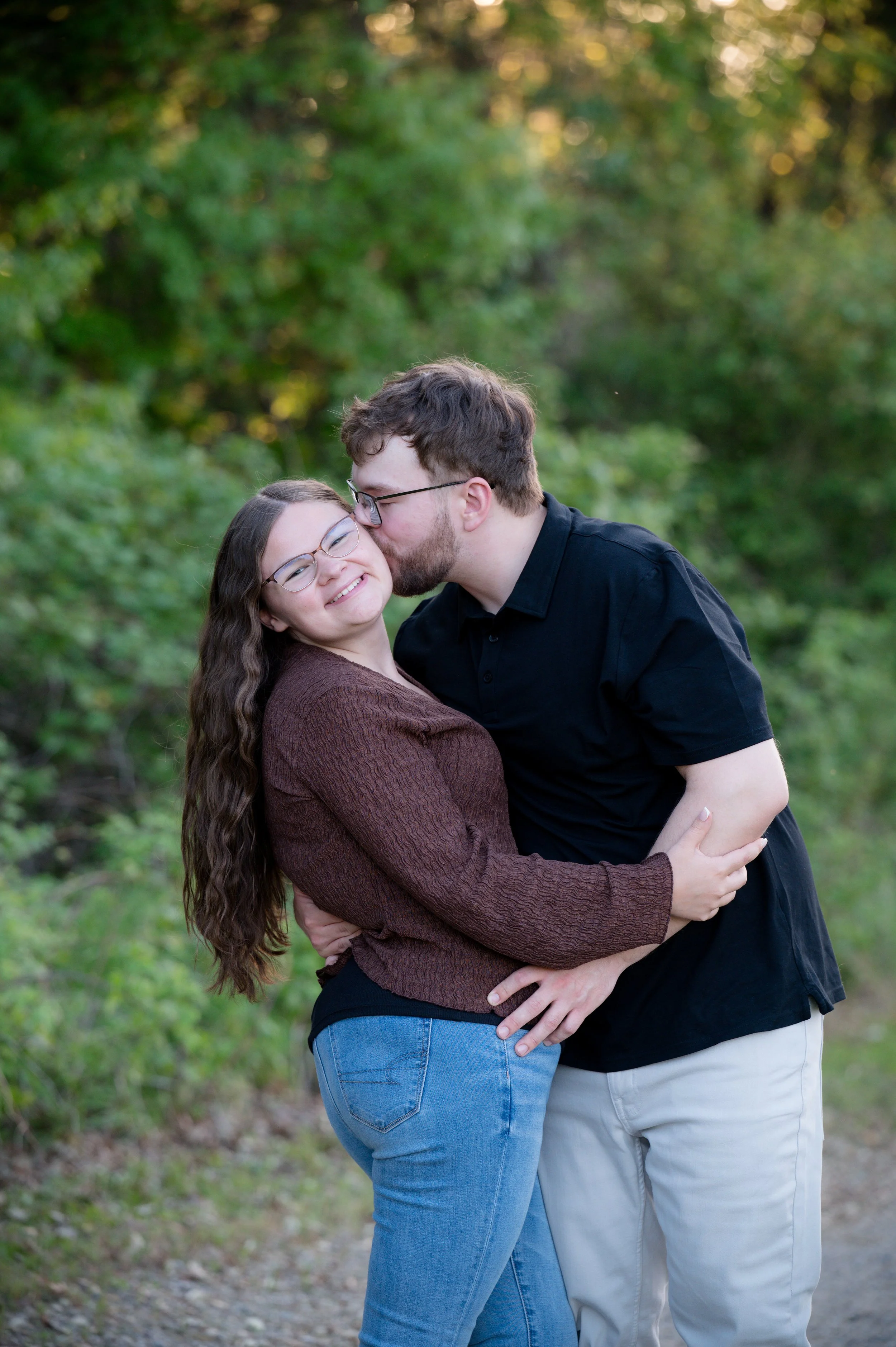 A couple hugging outdoors with a green, leafy background. The woman has long, curly brown hair and glasses, and the man has short, wavy brown hair and a beard. The man is kissing the woman on the cheek while she smiles.