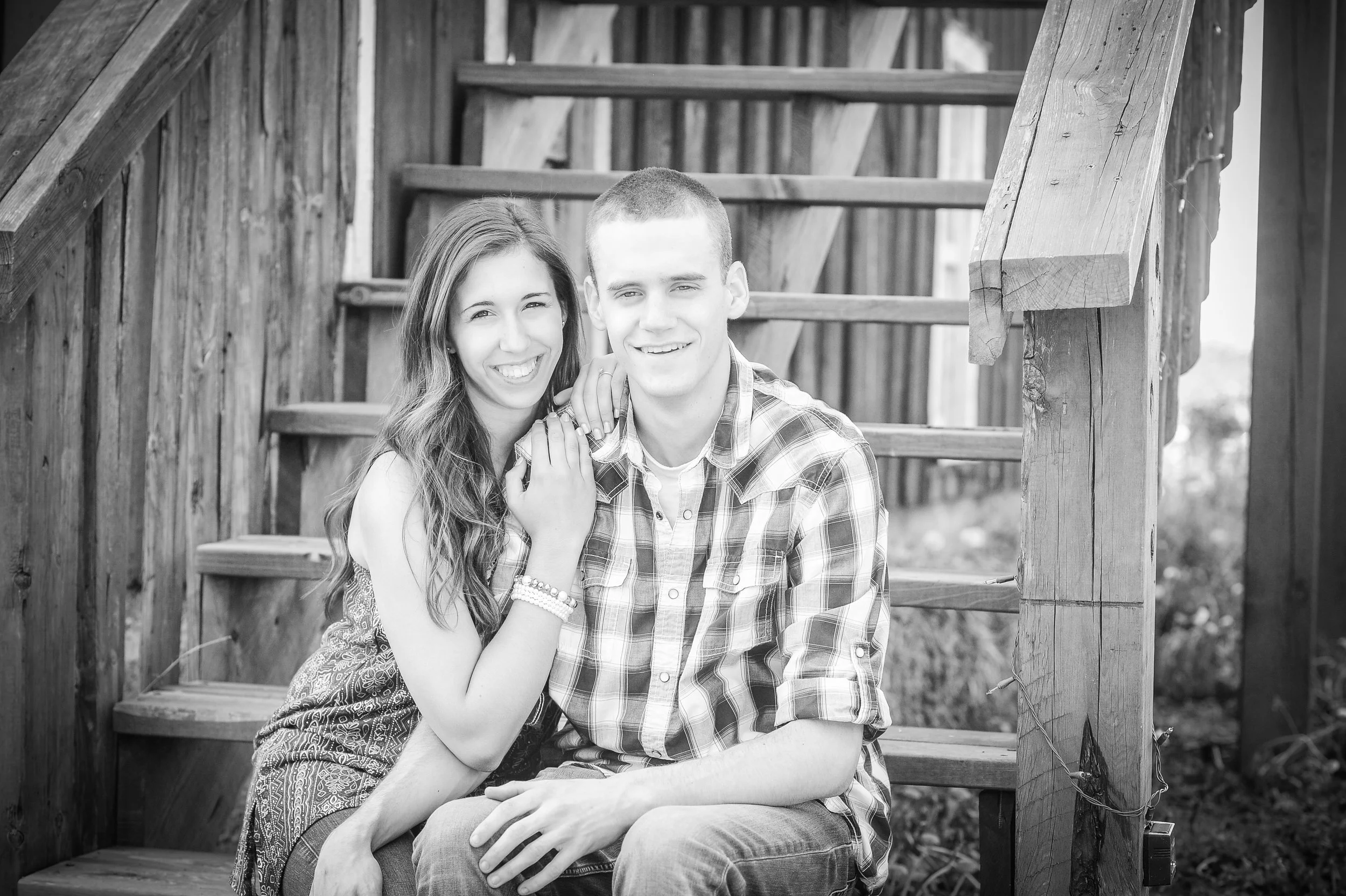 A black and white photo of a smiling young woman and young man sitting together on outdoor wooden stairs, with the woman affectionately resting her hand on the man’s shoulder.