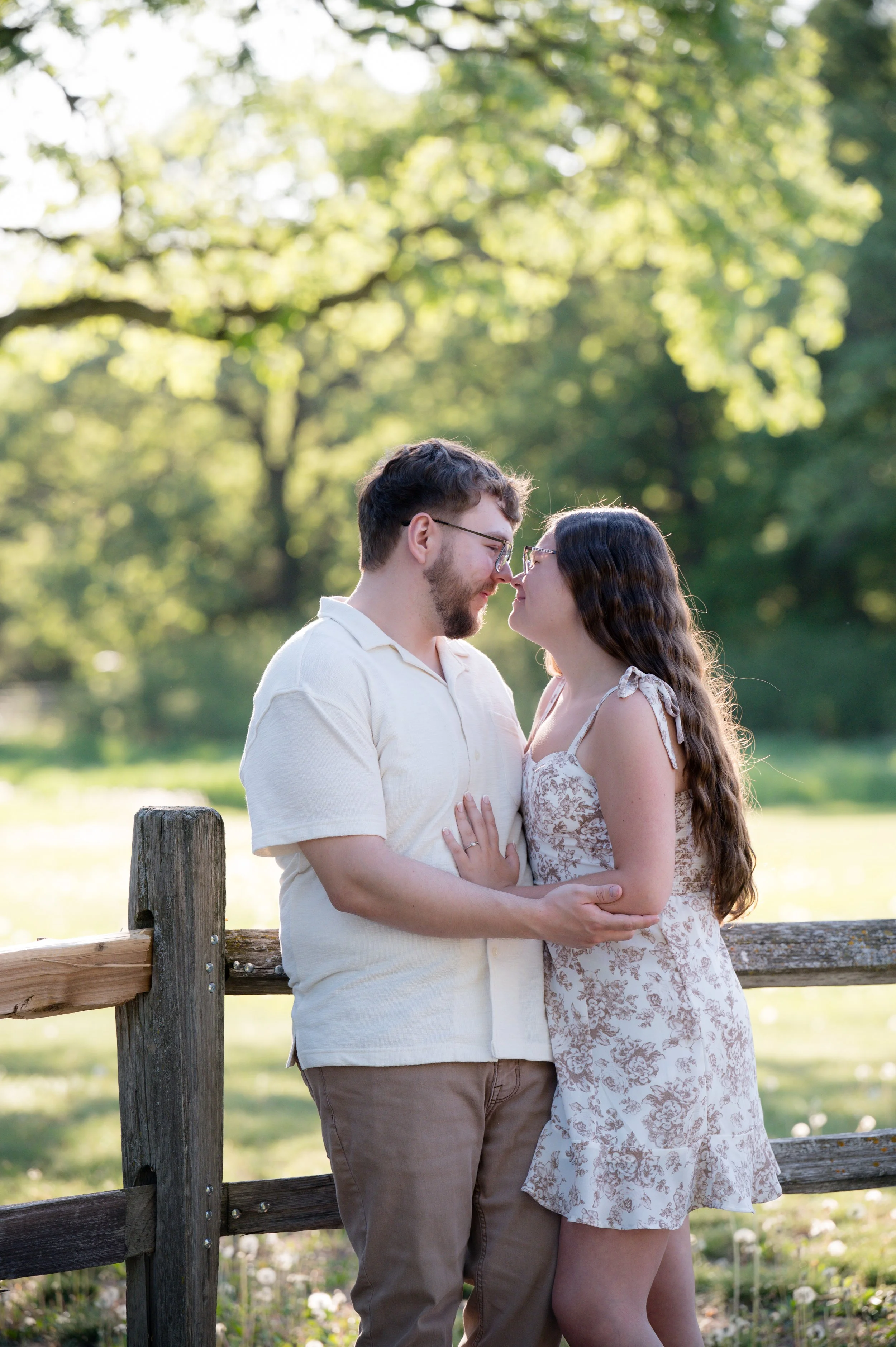 A young couple stands close together outdoors, touching foreheads and smiling, with a wooden fence and green trees in the background.