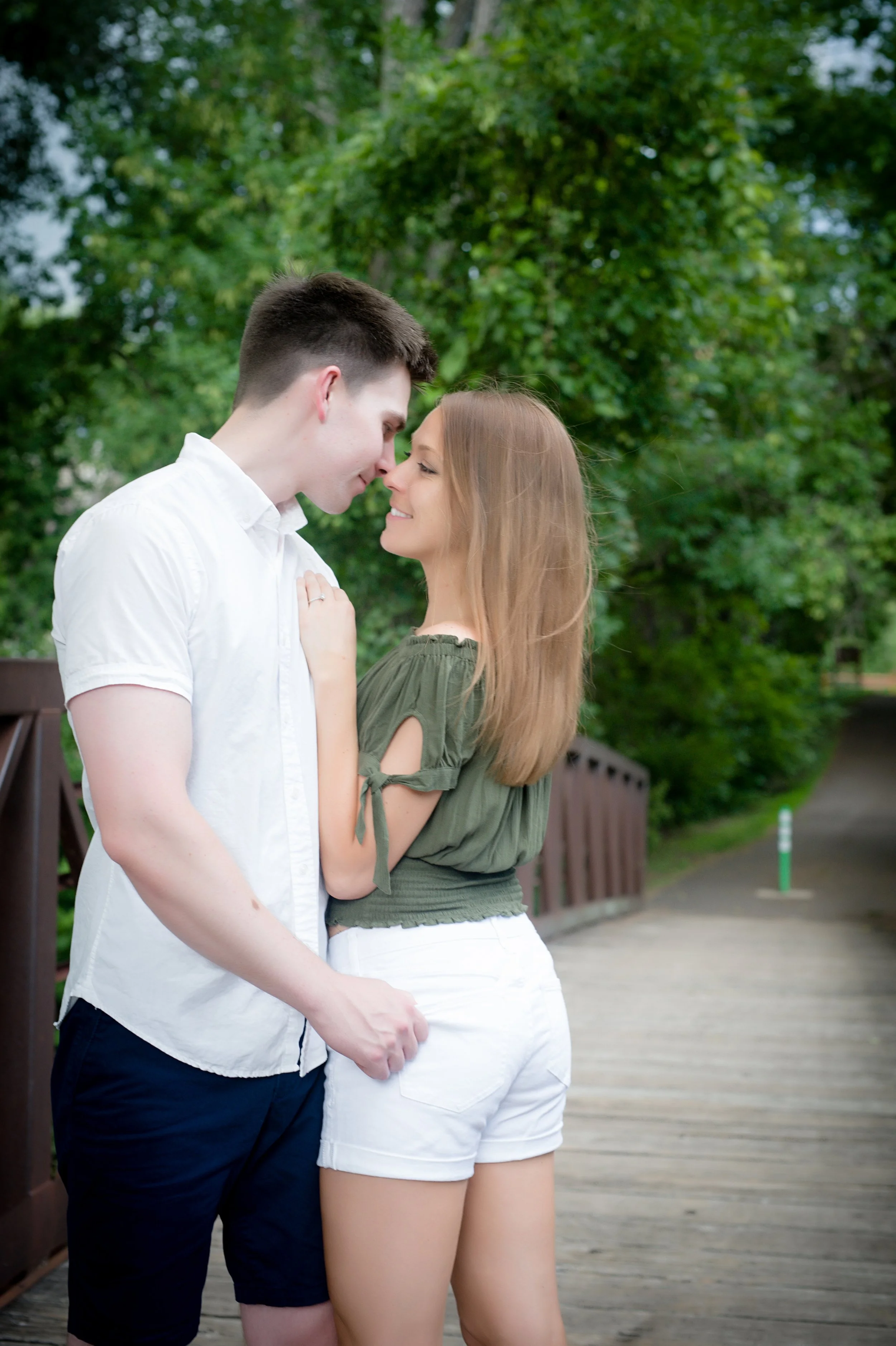 A young man and woman stand close to each other outdoors, touching foreheads and smiling. The man wears a white short-sleeve shirt and dark shorts, while the woman wears an olive-green top and white shorts. They are in a green, leafy park setting wit