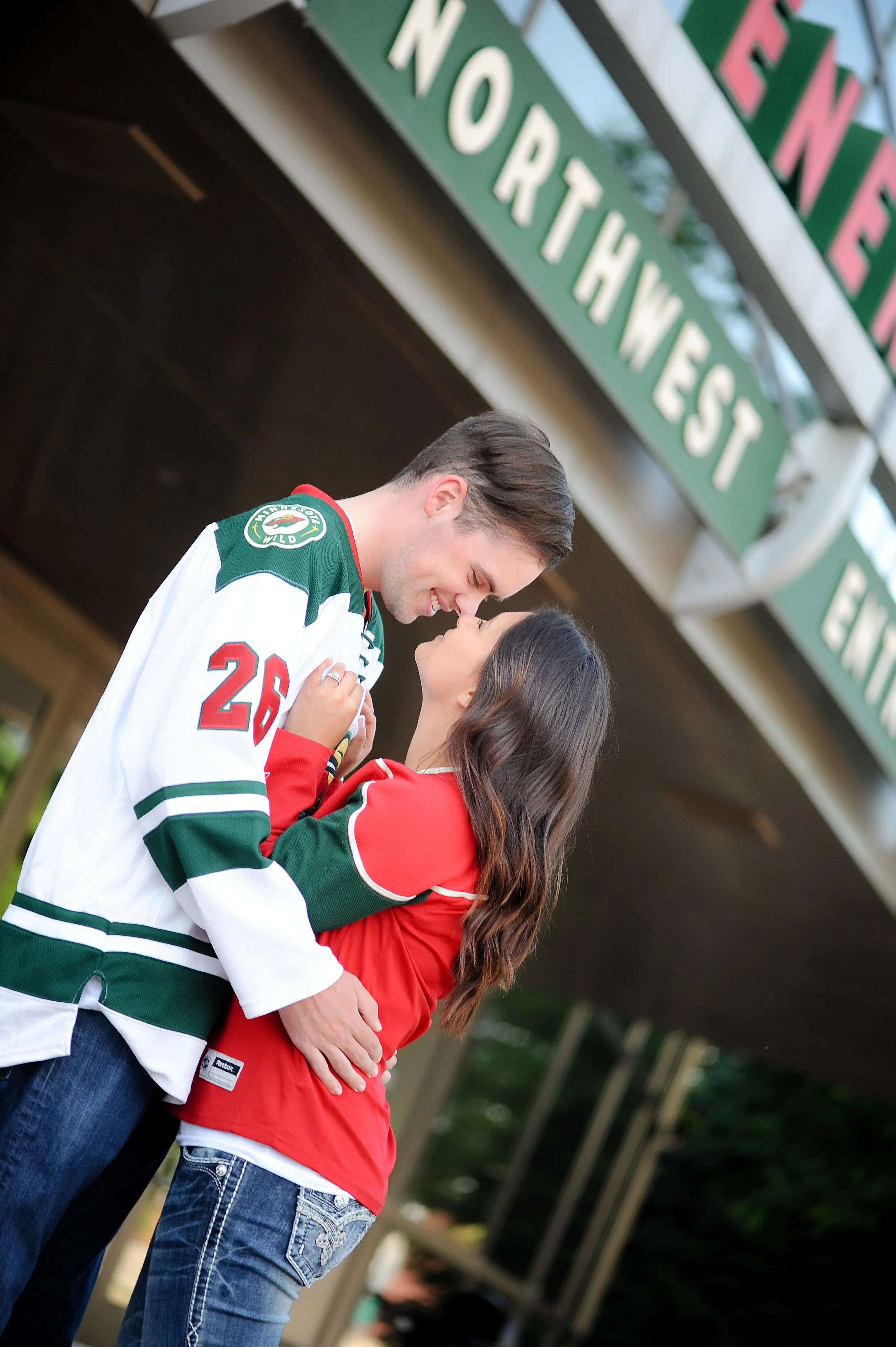 A young man and woman are standing close together, gazing into each other's eyes with happy expressions. The man is wearing a White Sox hockey jersey, and the woman is dressed in a red jacket and jeans. They are standing outdoors in front of a buildi
