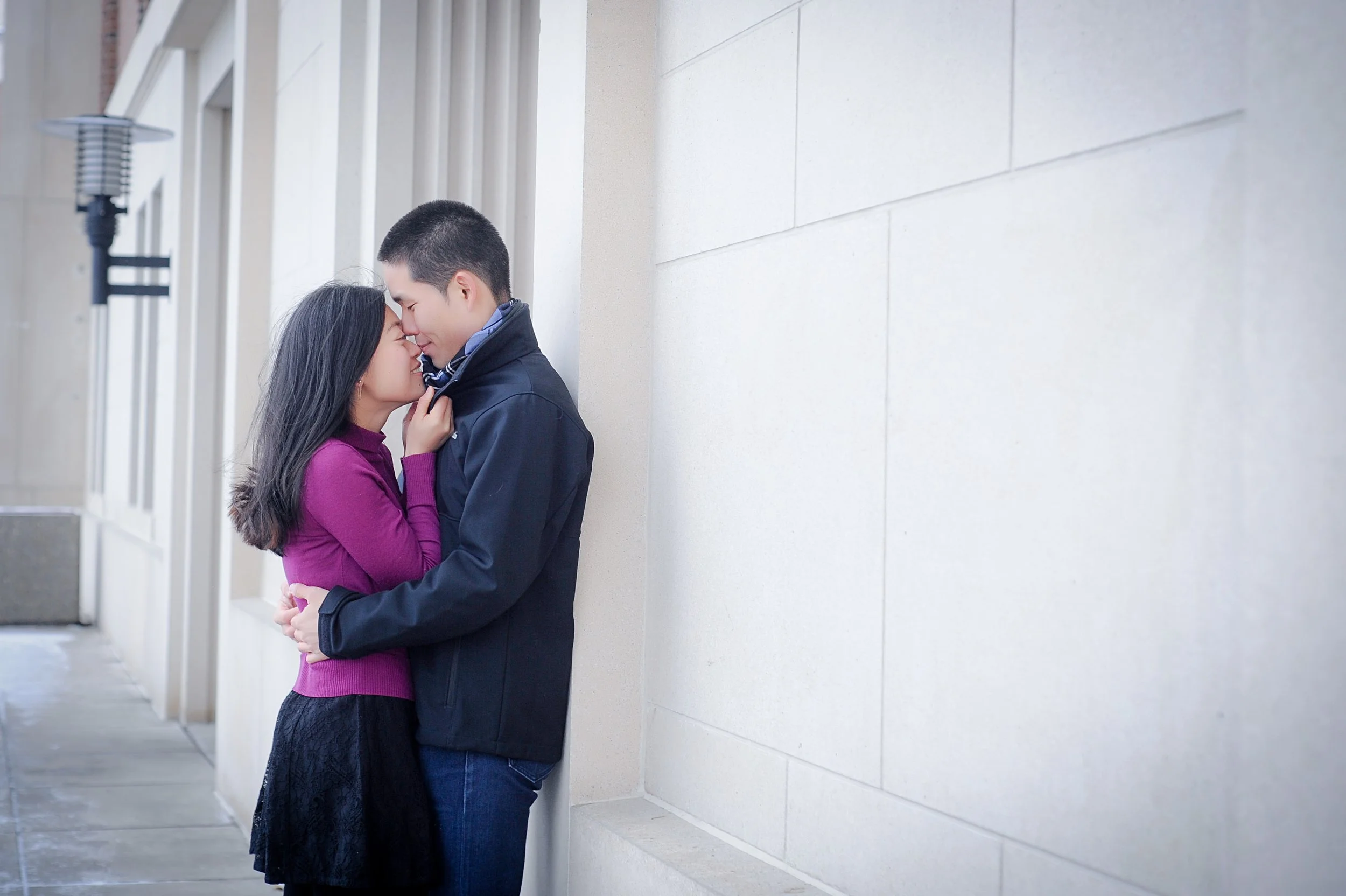 A young couple is standing close together, forehead to forehead, with their eyes closed, leaning against a pale-colored wall on the sidewalk in an urban setting.