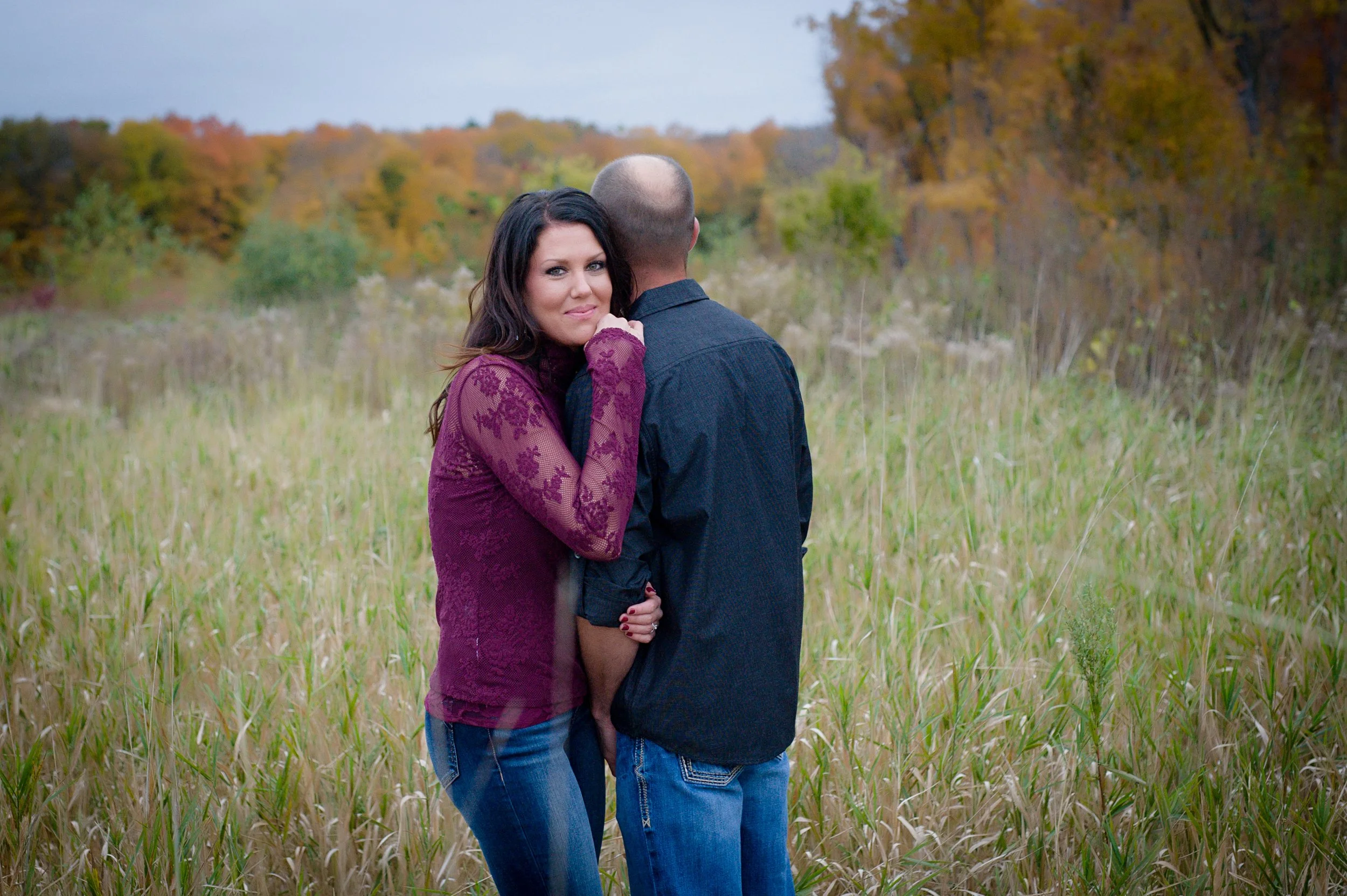 A woman with long dark hair wearing a burgundy lace top hugs a man with a shaved head wearing a dark shirt, standing in a field with tall grass and autumn trees in the background.