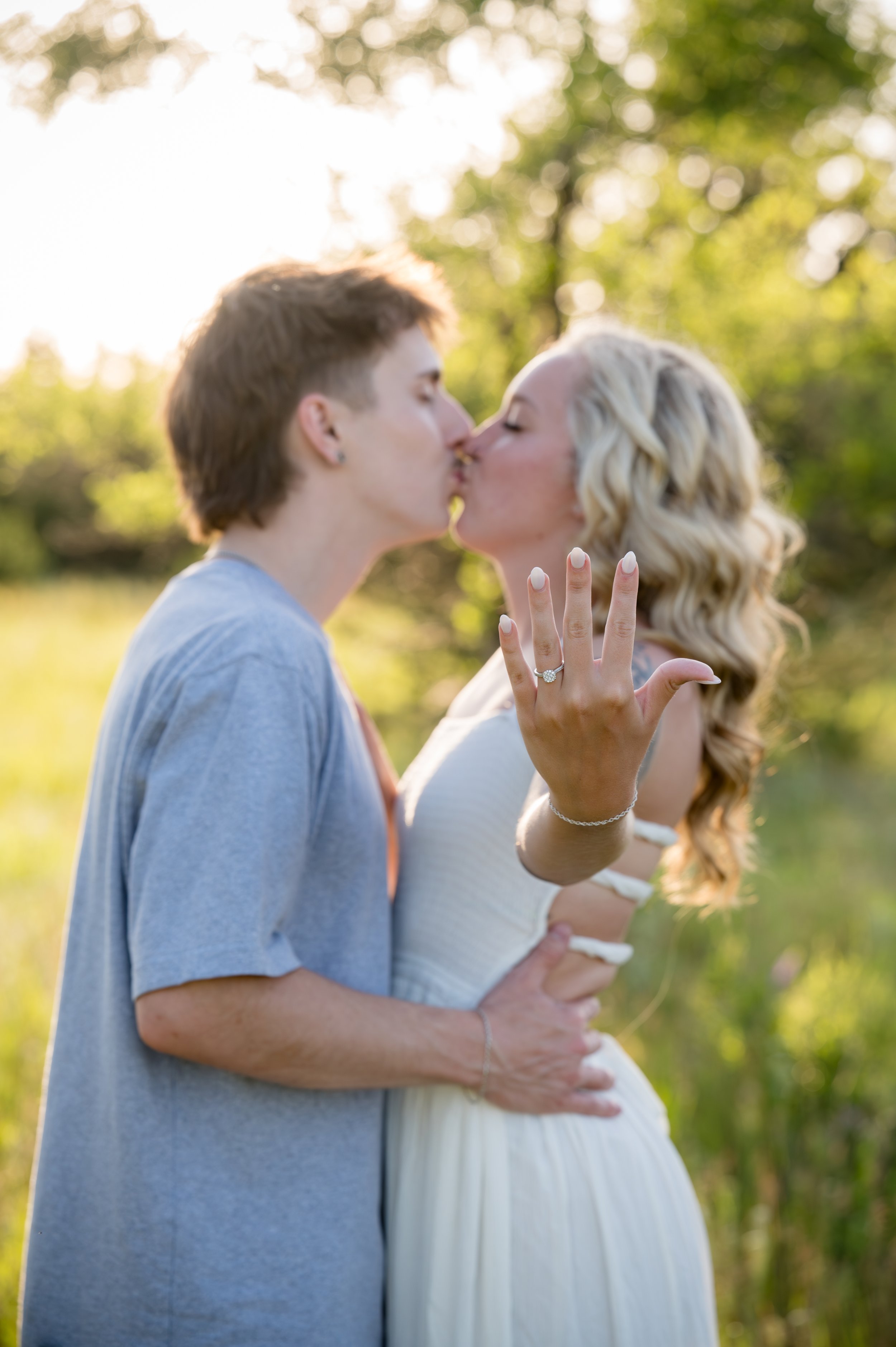 A man and a pregnant woman sharing a kiss outdoors during sunset, the woman showing her engagement ring and holding her baby bump.