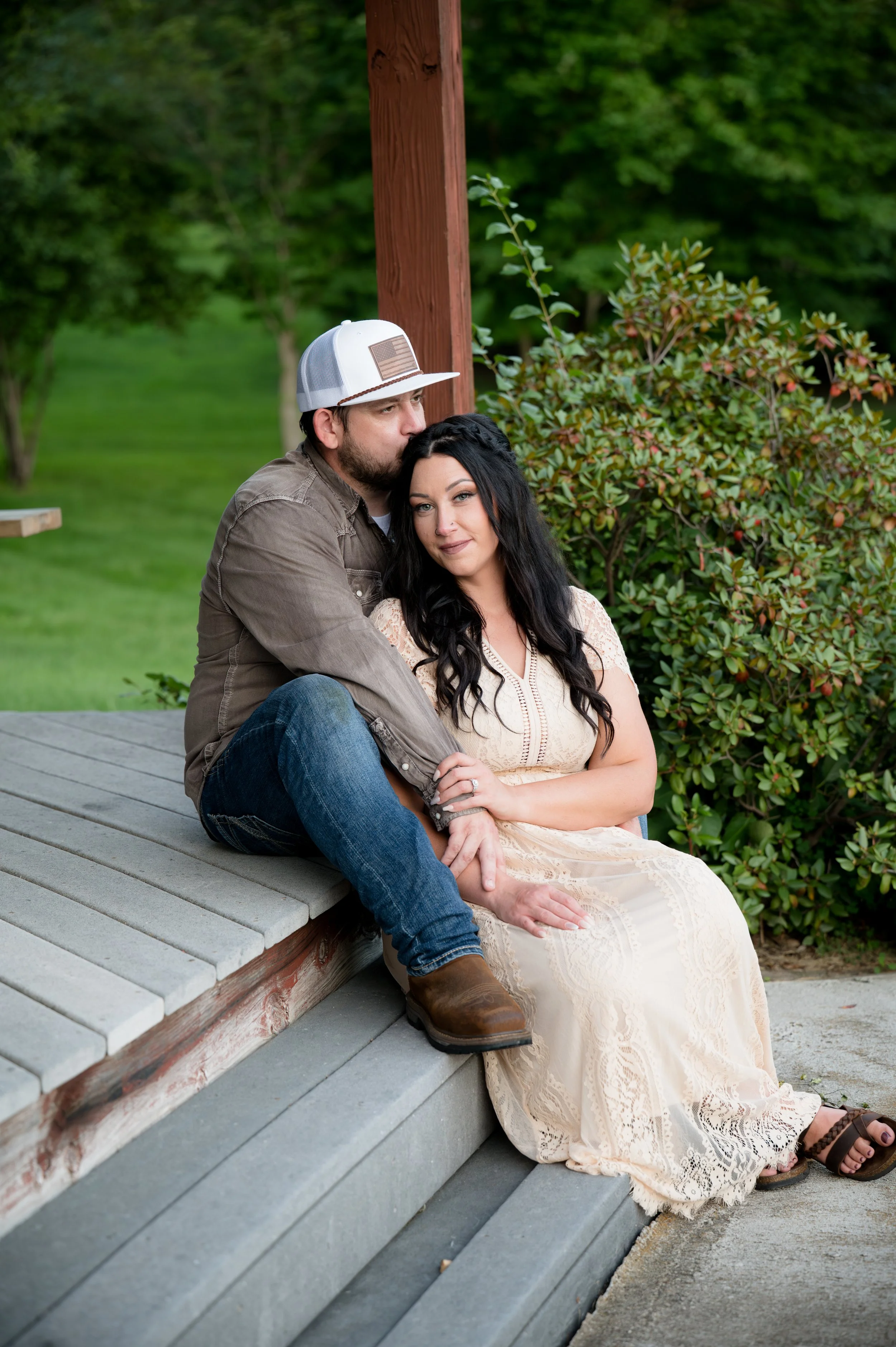 A couple sitting on outdoor steps, with the man kissing the woman's forehead, surrounded by green trees and bushes.