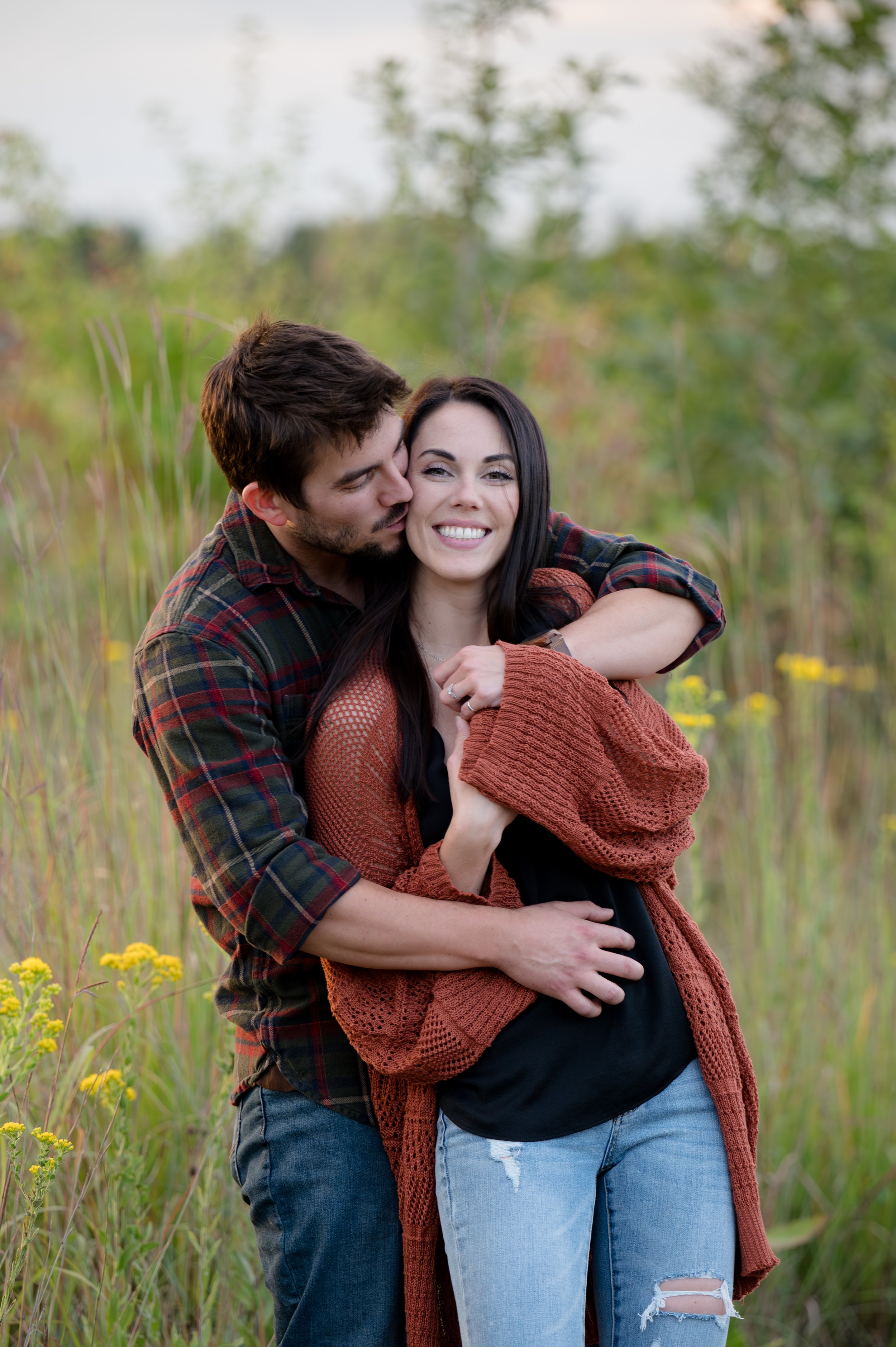 A young couple standing in a field of green grass and yellow flowers, embracing and smiling at the camera. The man is kissing the woman on the cheek, and they both look happy. The man has brown hair and a beard, wearing a plaid shirt. The woman has l