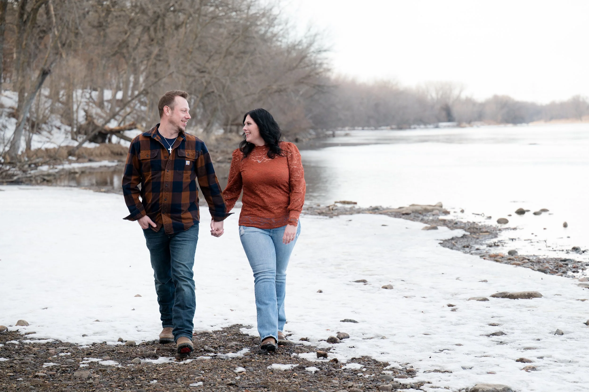 A couple walking hand in hand along a snow-covered riverbank with leafless trees in the background.