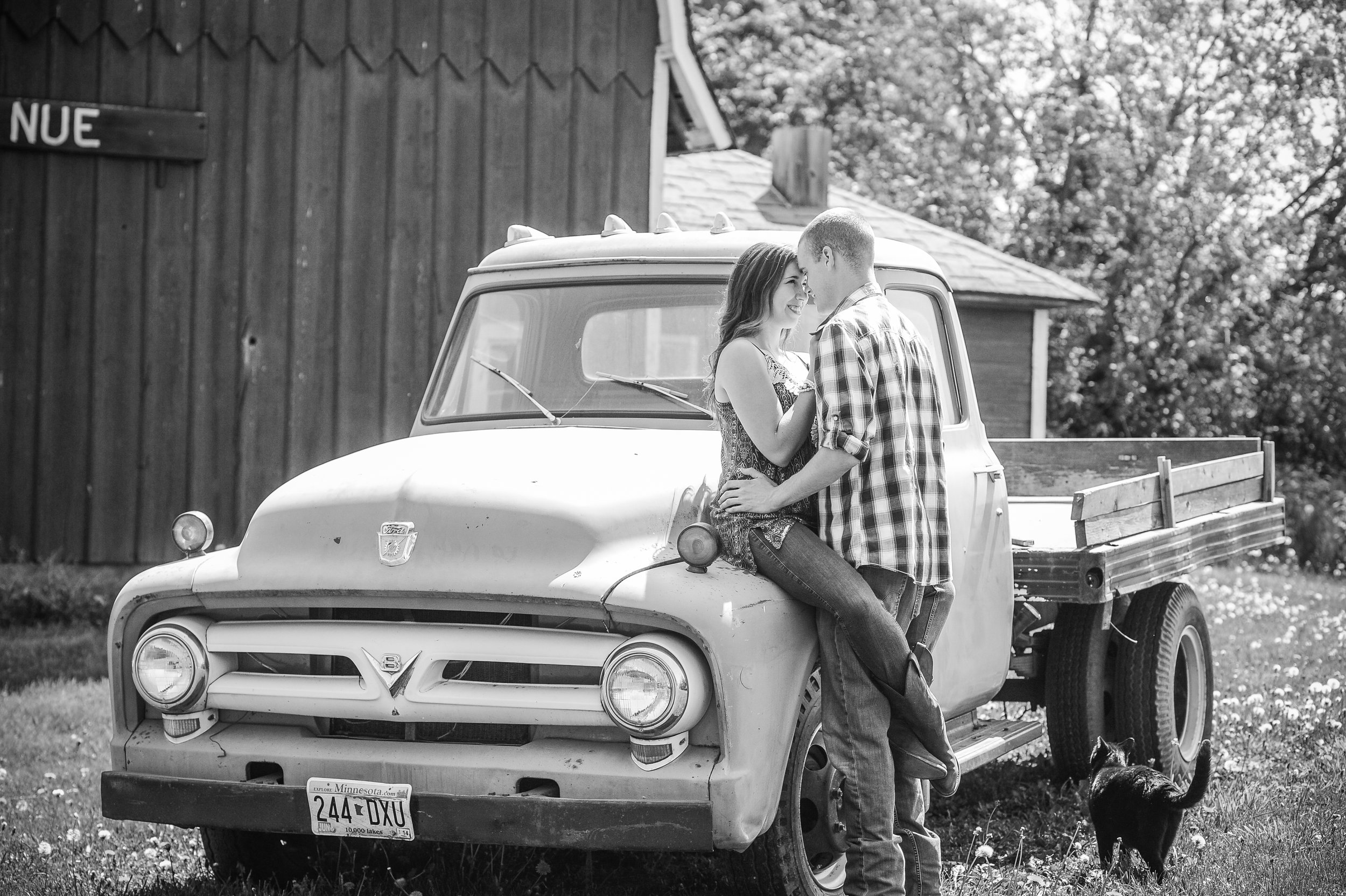 A couple stands close together on the hood of a vintage truck, smiling and gazing at each other. The woman is sitting on the truck, while the man is standing. There is a dog sitting on the ground nearby. The scene appears to be outdoors next to a woo