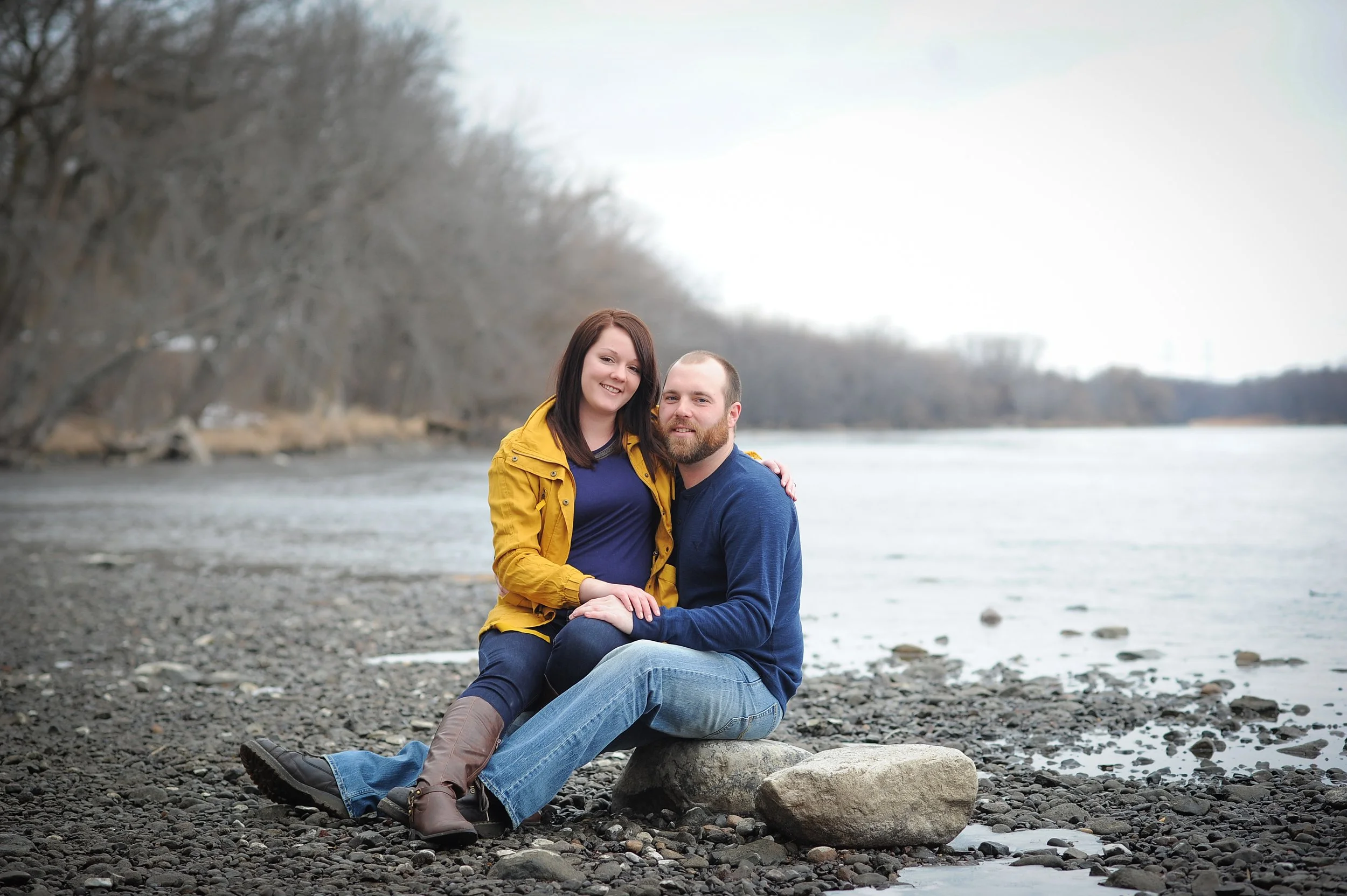 A smiling man and woman sitting on a riverbank with trees in the background and water behind them.