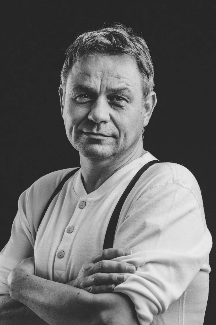 Ett svart-hvitt portrett av Geir Magne, som står med armene krysset mot en mørk bakgrunn.
