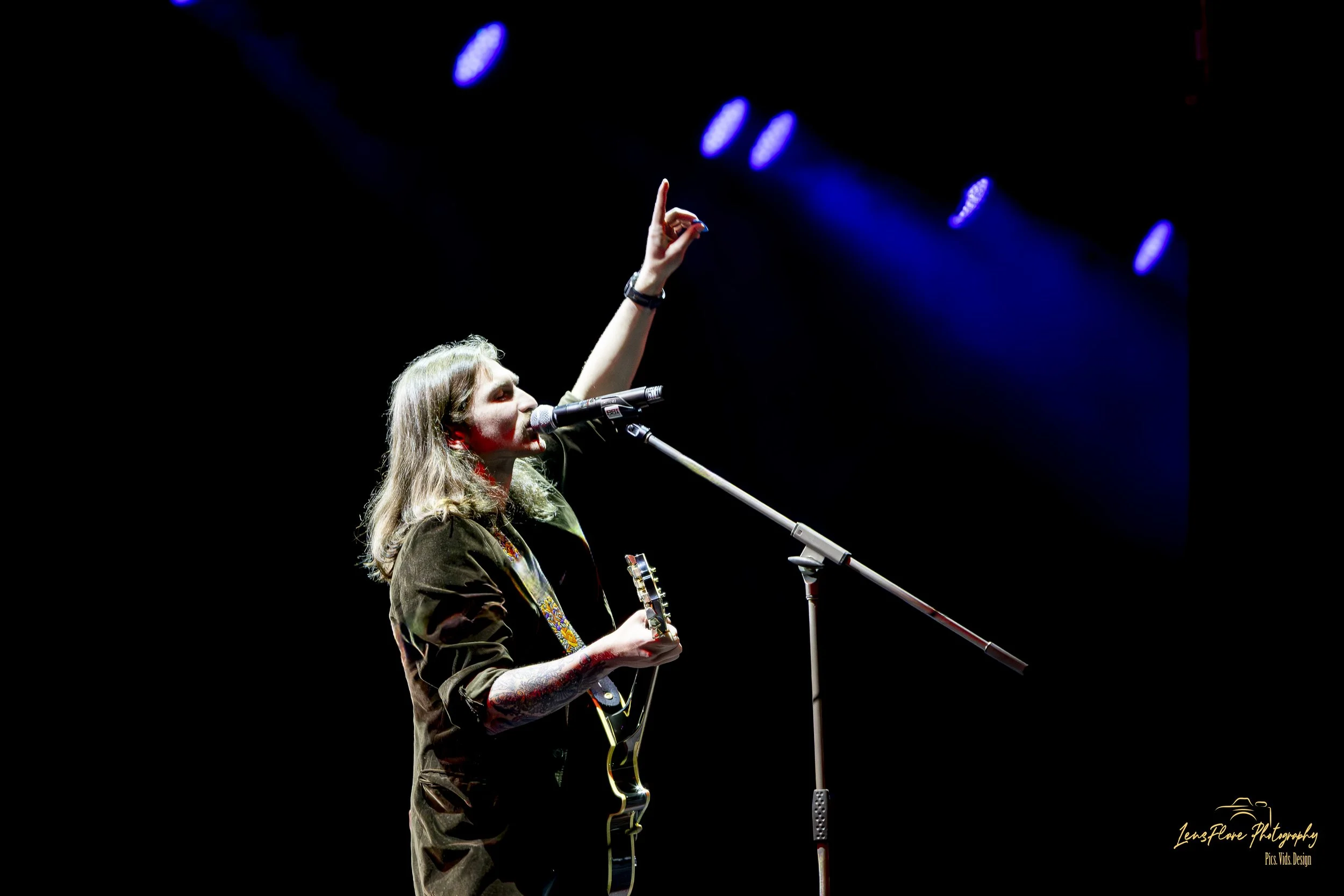 A woman with long hair, tattoos, and a guitar, singing into a microphone on stage, raising her hand upward against a dark background with blue stage lights.