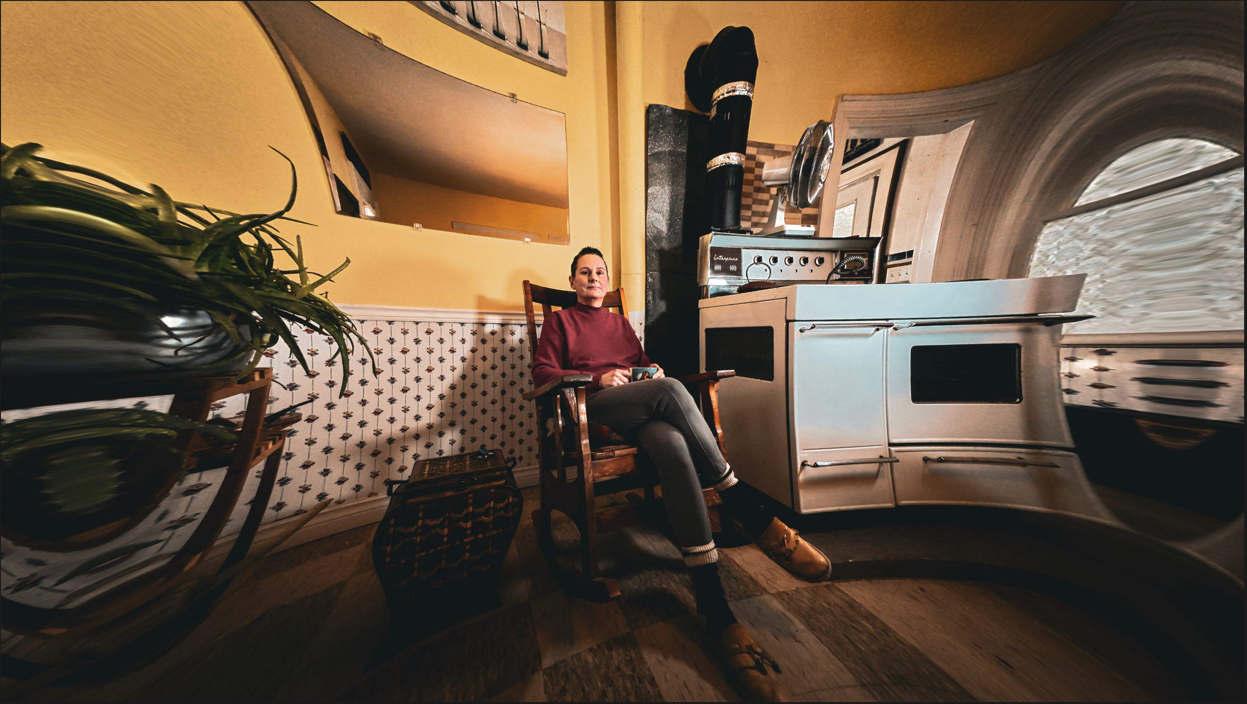 A woman sitting on a wooden chair in a kitchen, holding a cup. The kitchen has yellow walls, a white stove, and a window. There is a plant on a stand to the left and a suitcase on the floor in front of her. The photo has a distorted, curved perspecti