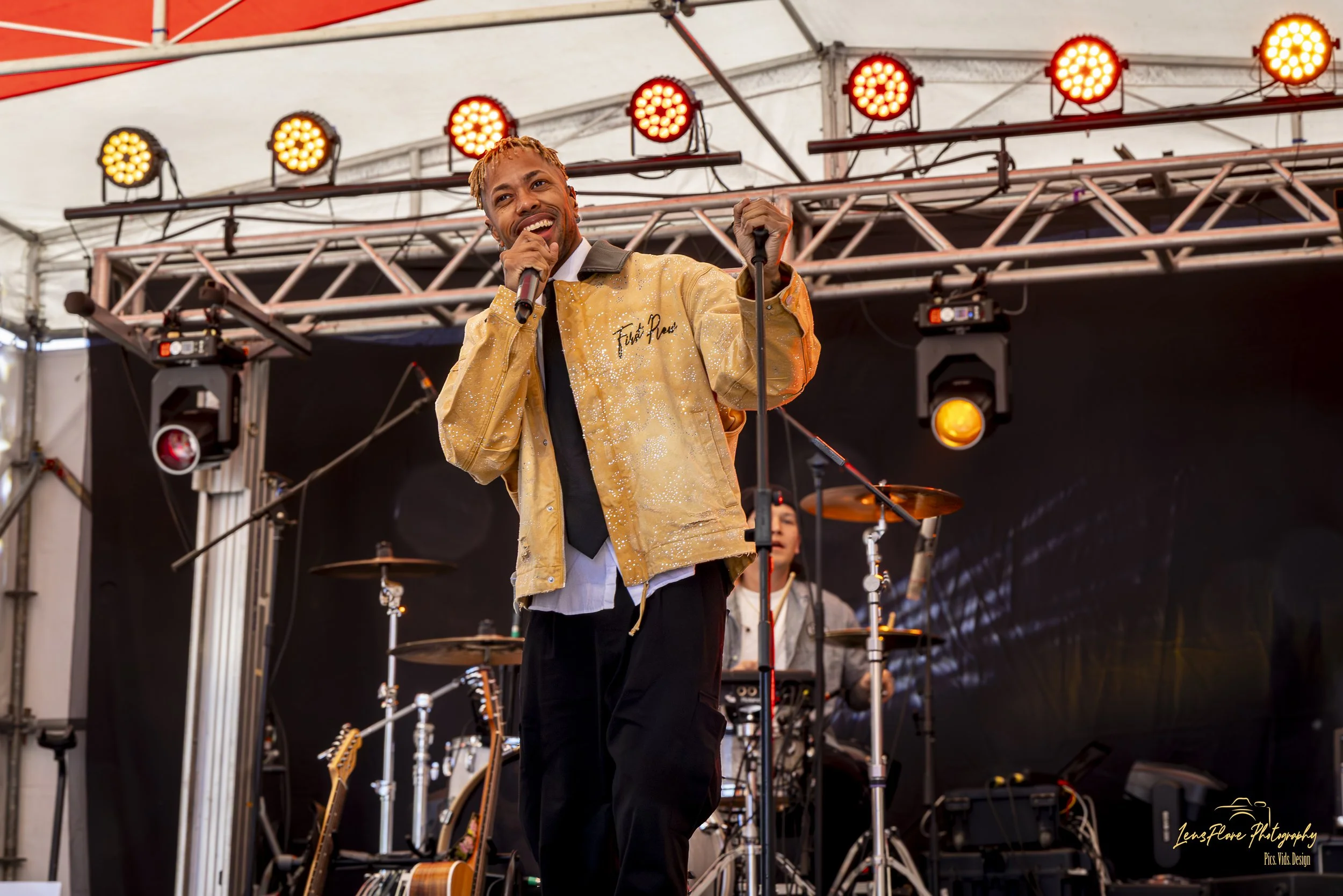 Male singer performing on stage at an outdoor event, surrounded by musical instruments and stage lighting.