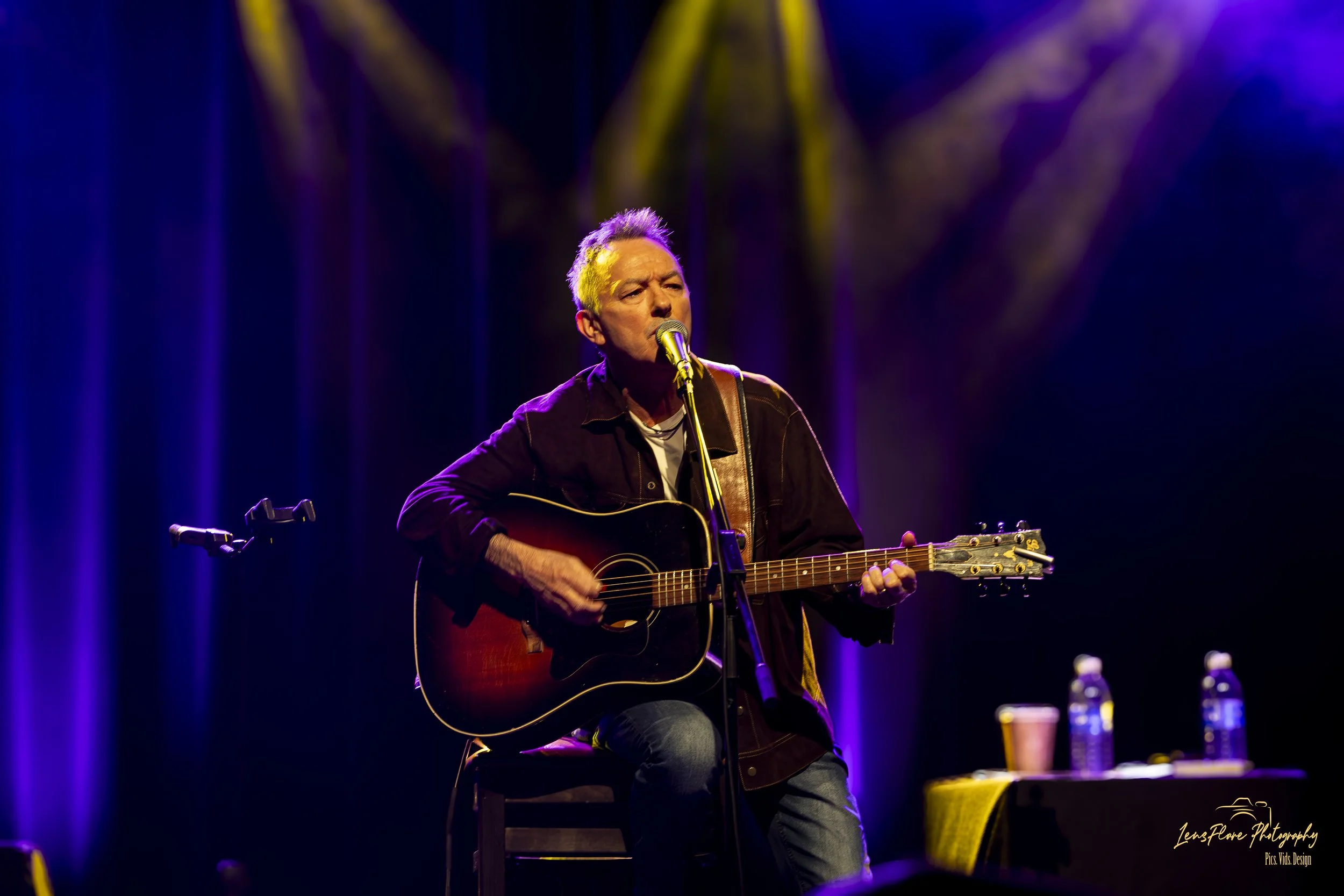 A man with gray hair playing an acoustic guitar and singing into a microphone on a stage with purple and yellow lighting.