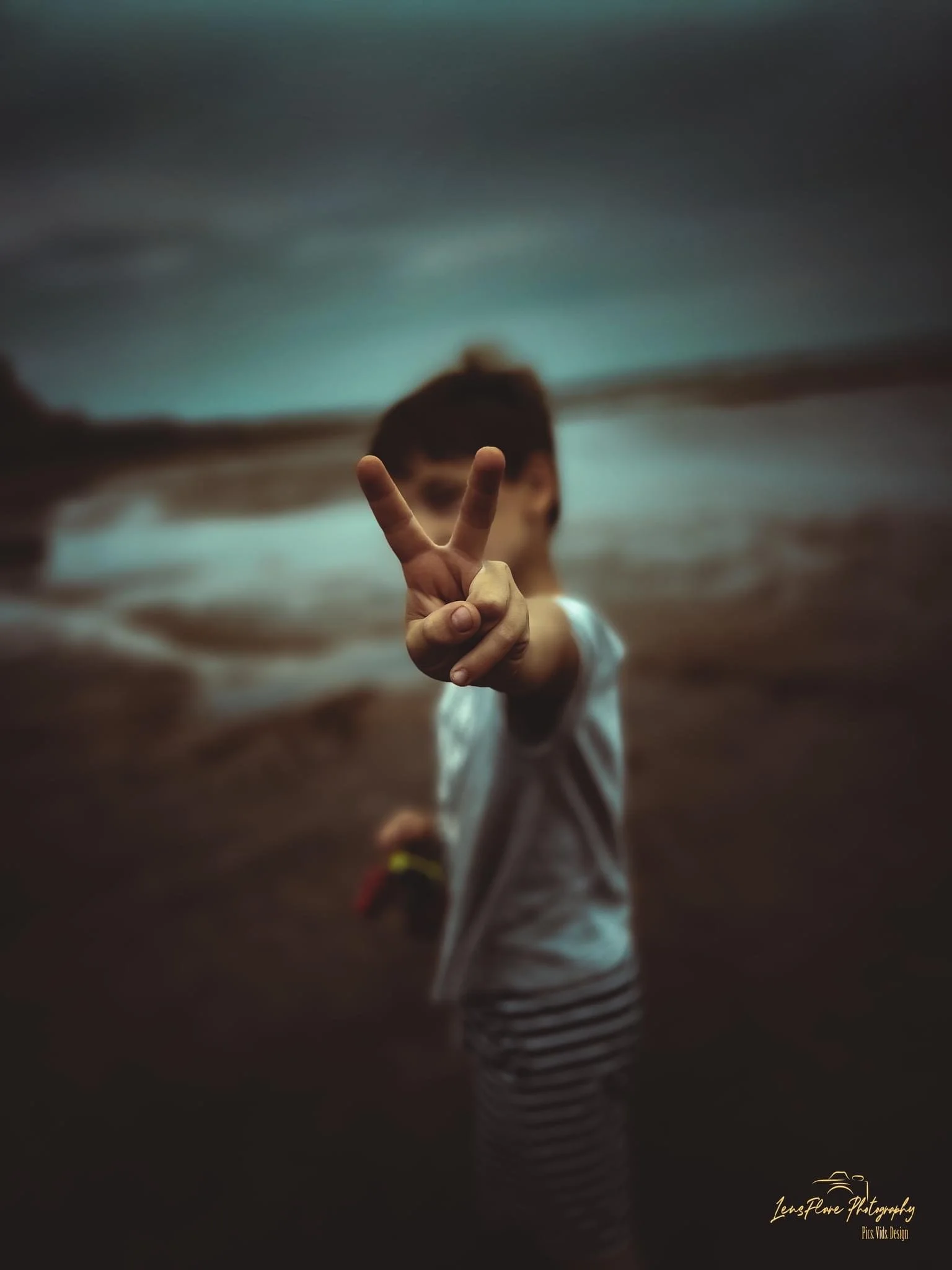 A young girl standing on a beach at dusk, holding up her hand in a peace sign towards the camera, with her face blurred in the background.