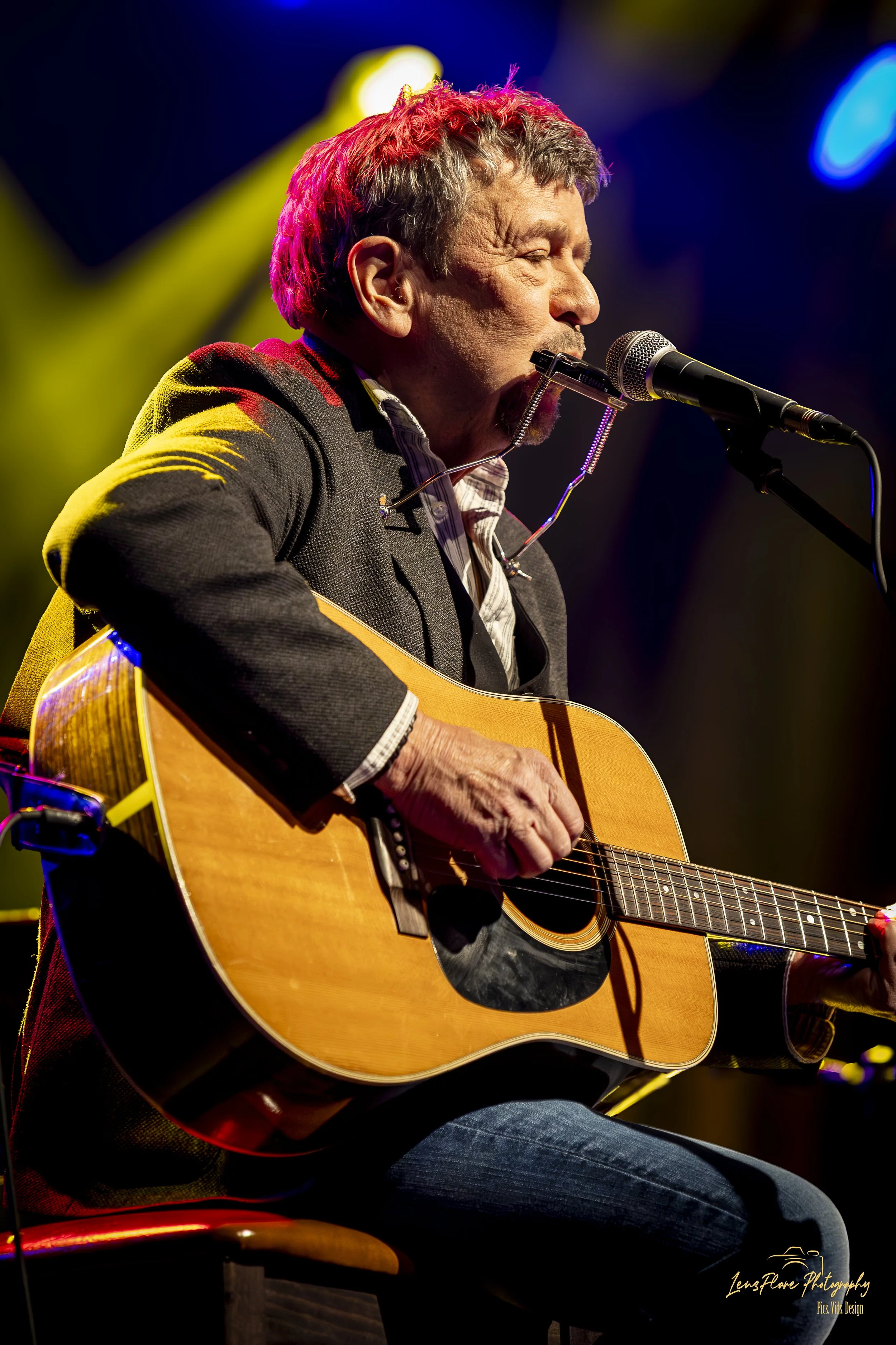 A man with short gray hair and a beard, wearing a gray blazer over a plaid shirt, is playing an acoustic guitar and singing into a microphone on a stage with colorful lights.