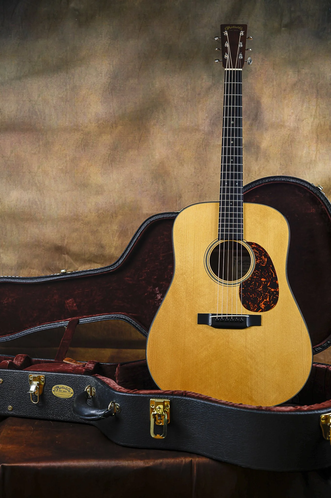 An acoustic Martin guitar with a natural wood finish, black pickguard, and six strings, resting inside a red velvet-lined black guitar case with gold latches, on a wooden surface.