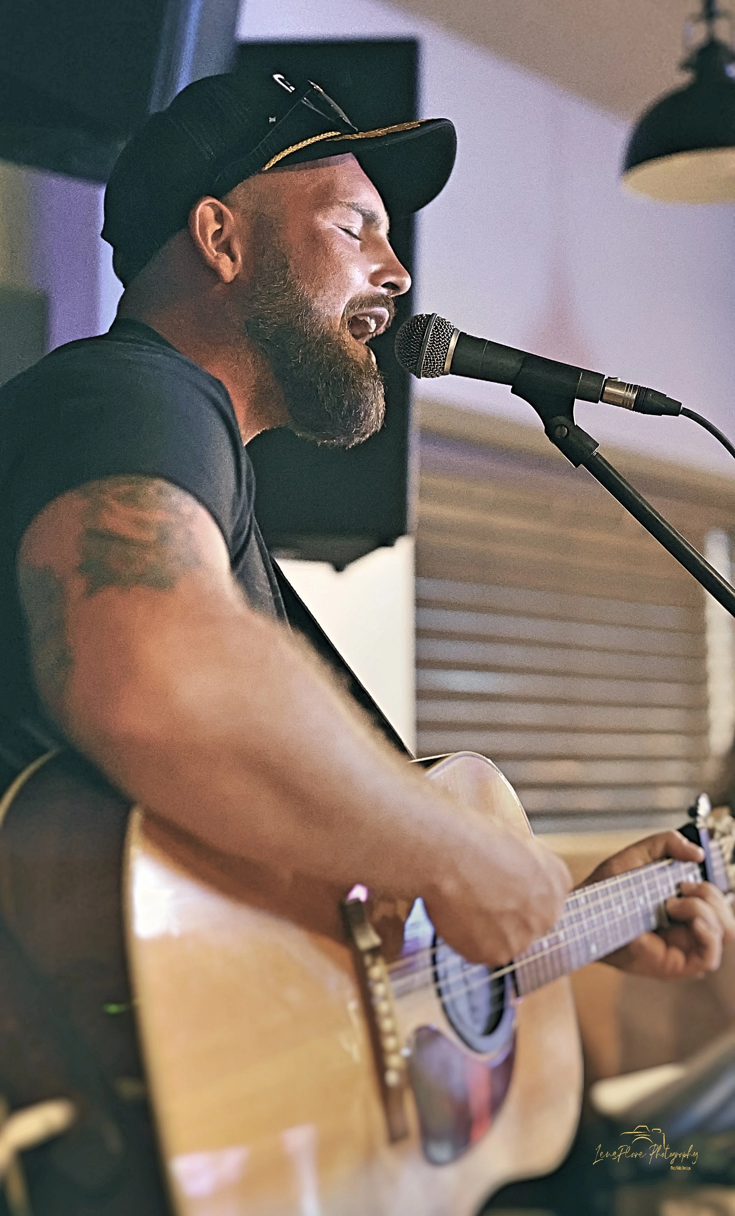A man with a beard singing into a microphone while playing an acoustic guitar, wearing a black cap and sunglasses on his head, inside a cozy indoor setting.