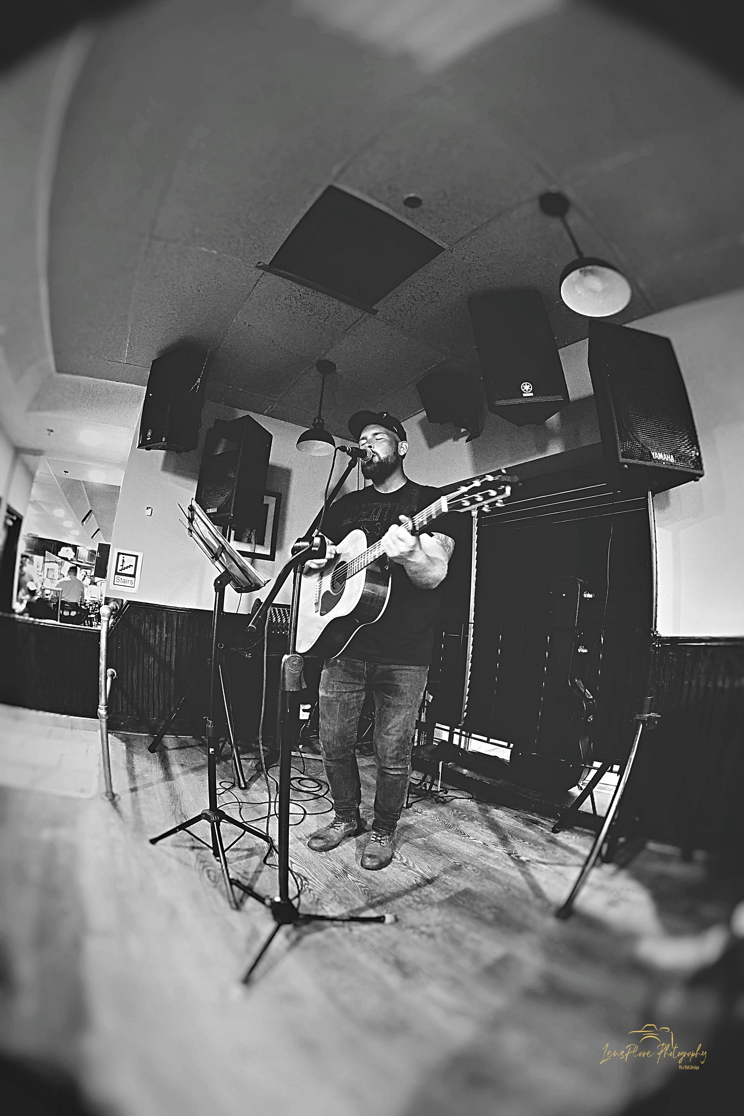 A man performing with an acoustic guitar on stage, singing into a microphone in a casual indoor setting, with audio speakers and music equipment around him.