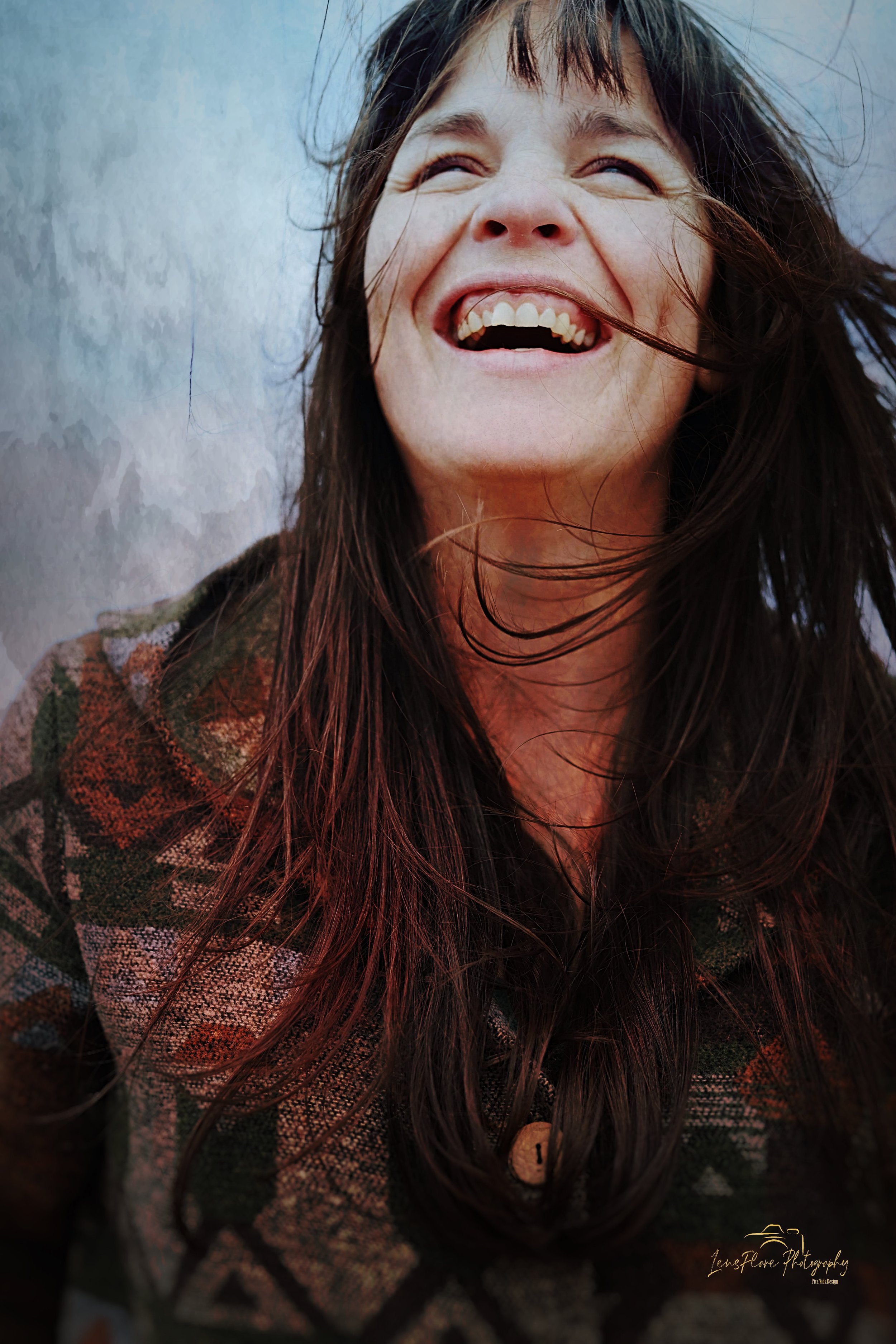 A woman with long dark hair is smiling and looking up, with her hair slightly blowing in the wind, against a cloudy sky.